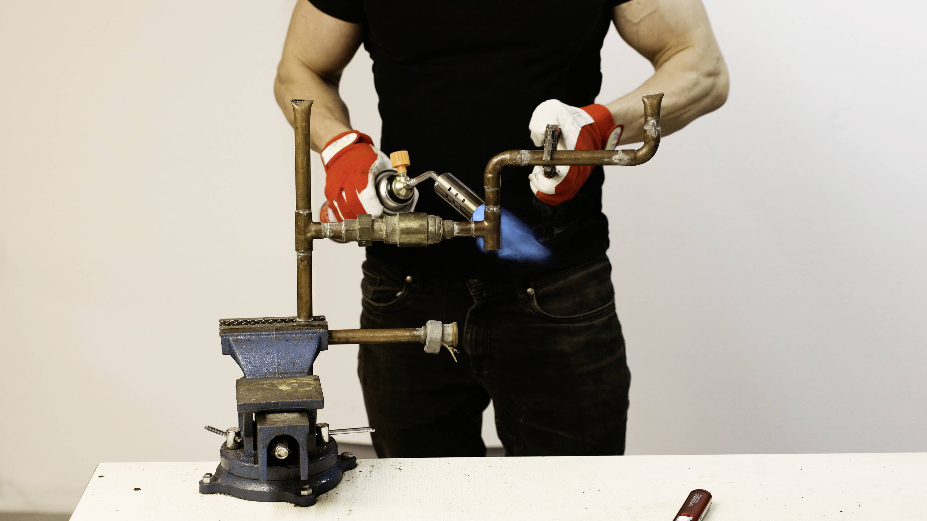 A person is working with a blowtorch on a metal pipe system clamped in a vice. They are wearing red gloves.