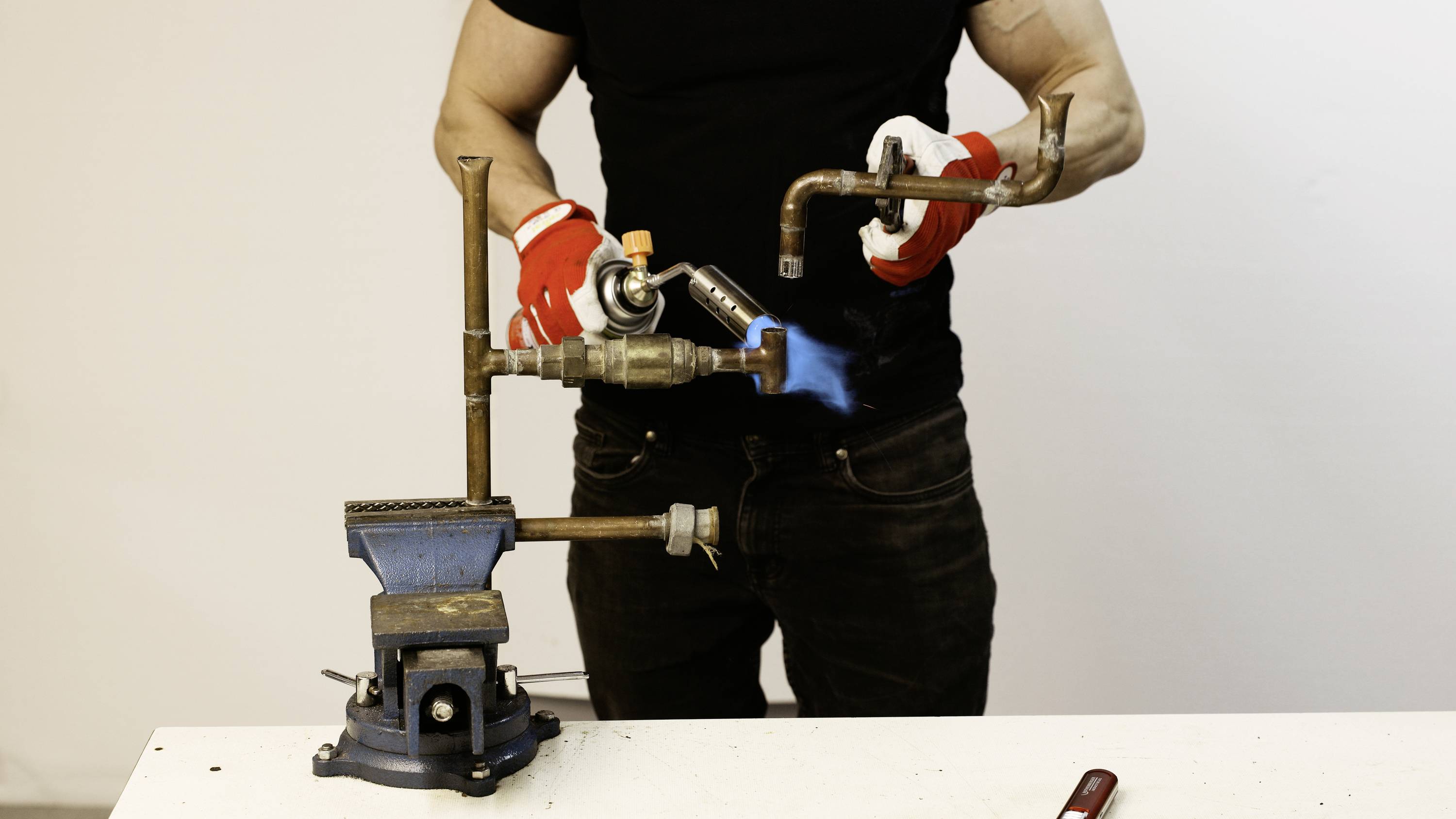A person wearing black clothing is using a blowtorch to heat metal pipes clamped in a vice.
