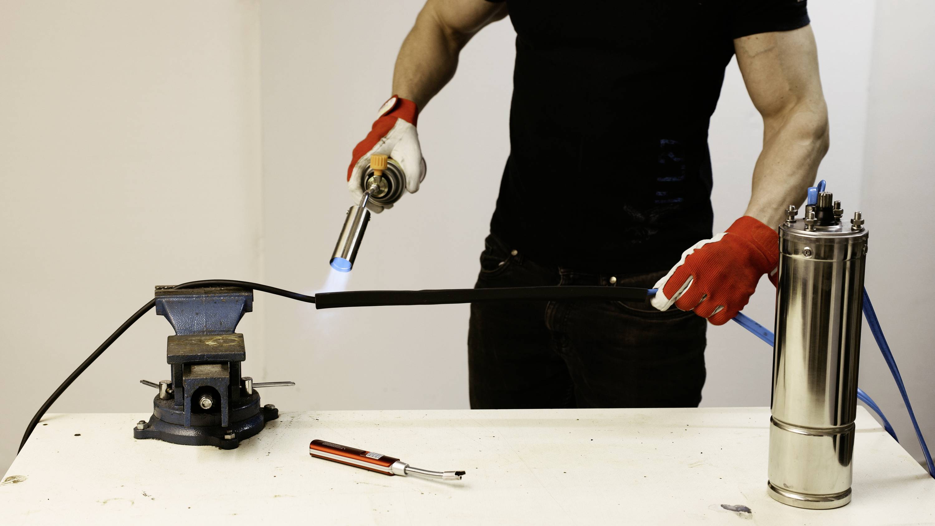 A person is working on a metal piece on a table using a gas flame. A metallic container is positioned to the right; appropriate red protective gloves are being worn.