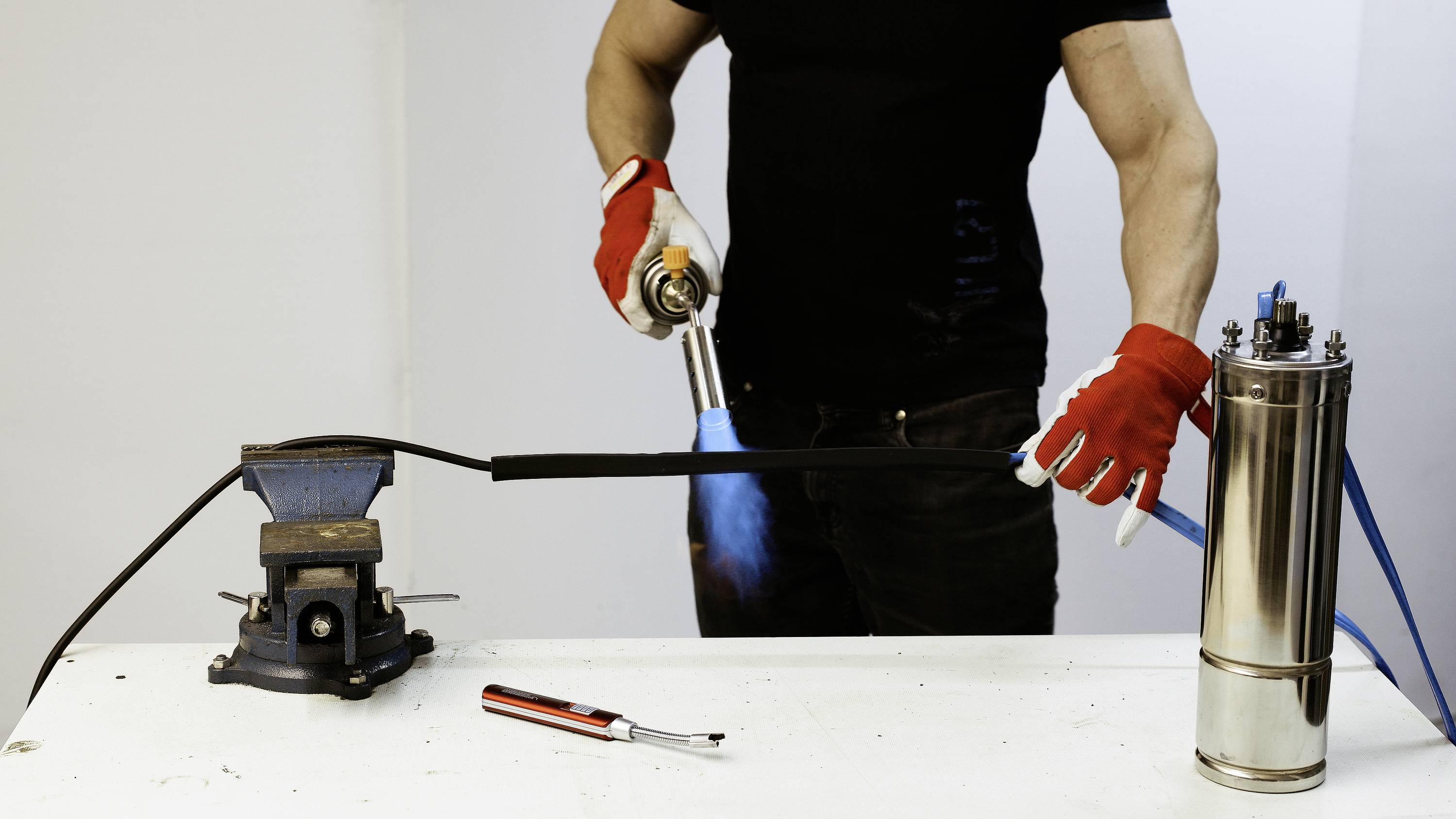 A person wearing a black T-shirt and red gloves is heating a black pipe clamped in a vice using a blowtorch.