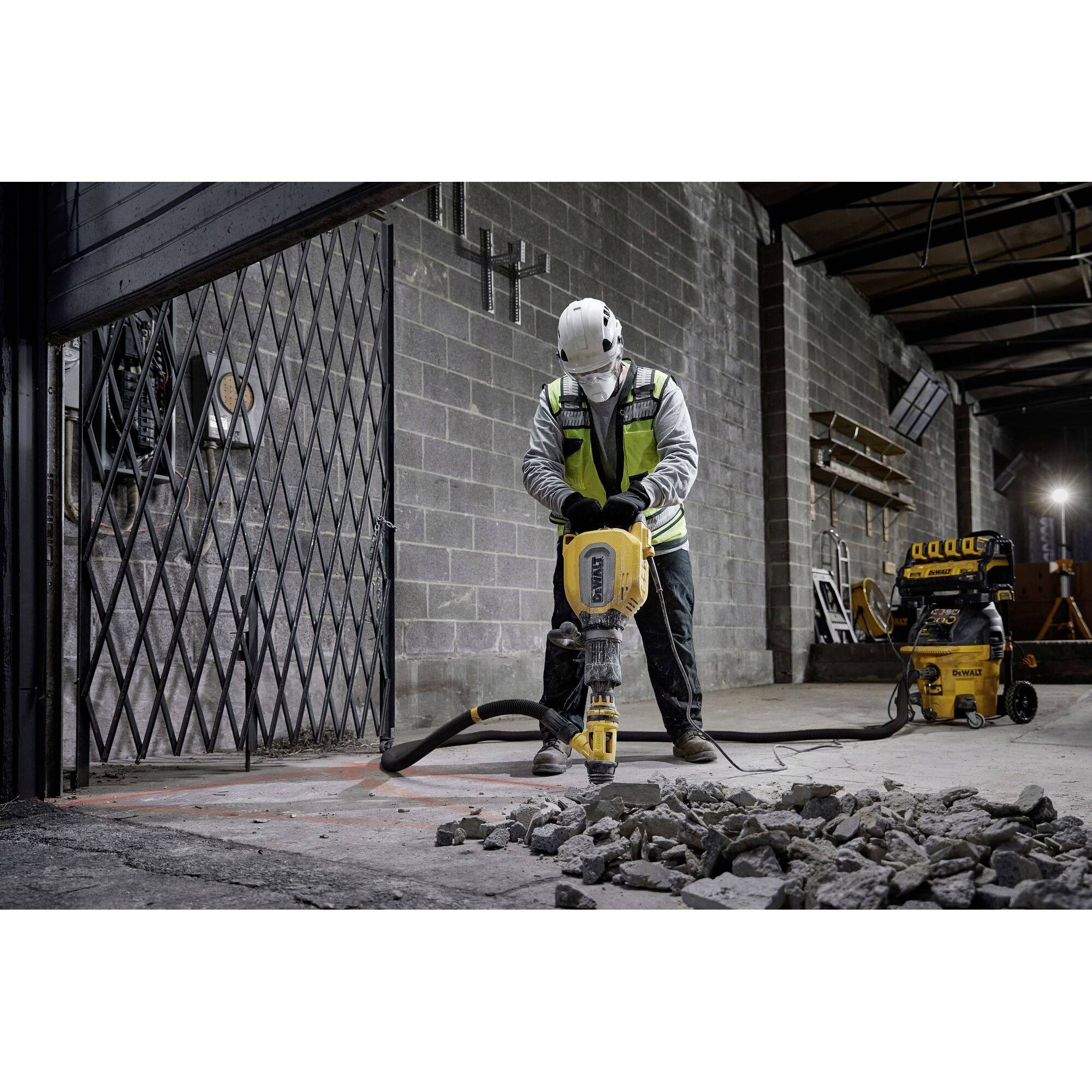 A construction worker wearing a hard hat and high-visibility vest is using a pneumatic drill to break up a concrete floor in an industrial setting.