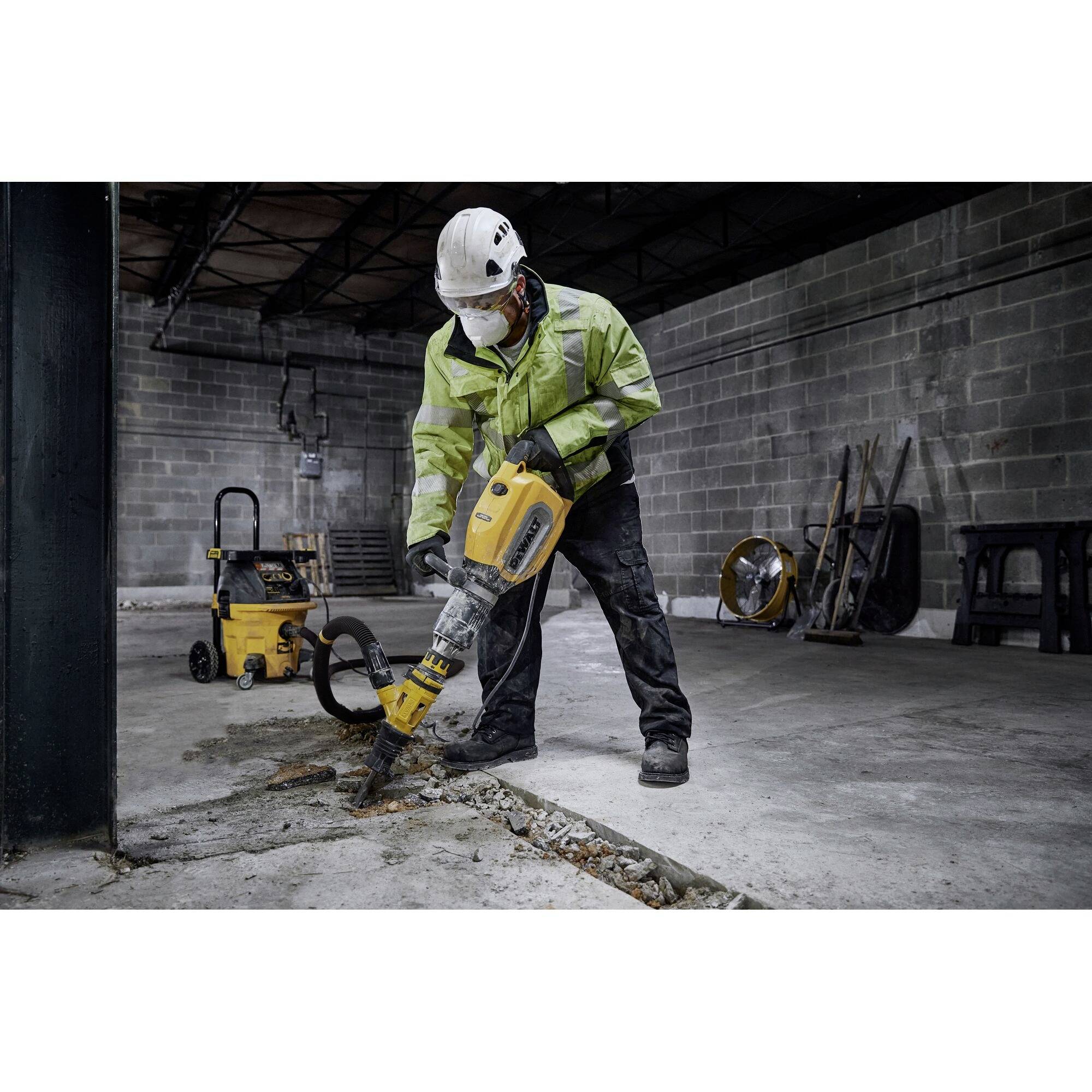 A worker wearing a safety helmet and protective clothing is using a pneumatic hammer to break up a concrete floor in a workshop. Tools are visible in the background.