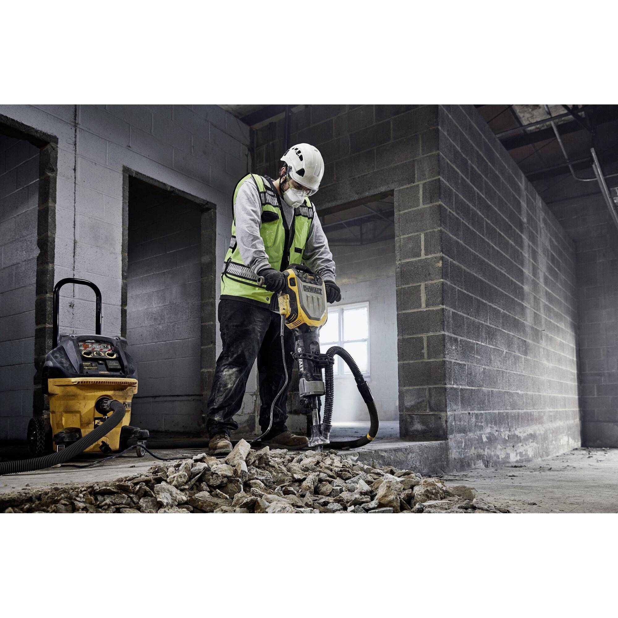 A construction worker wearing protective clothing and a hard hat is using a pneumatic drill to break up concrete in an unfinished building. A vacuum cleaner stands nearby.