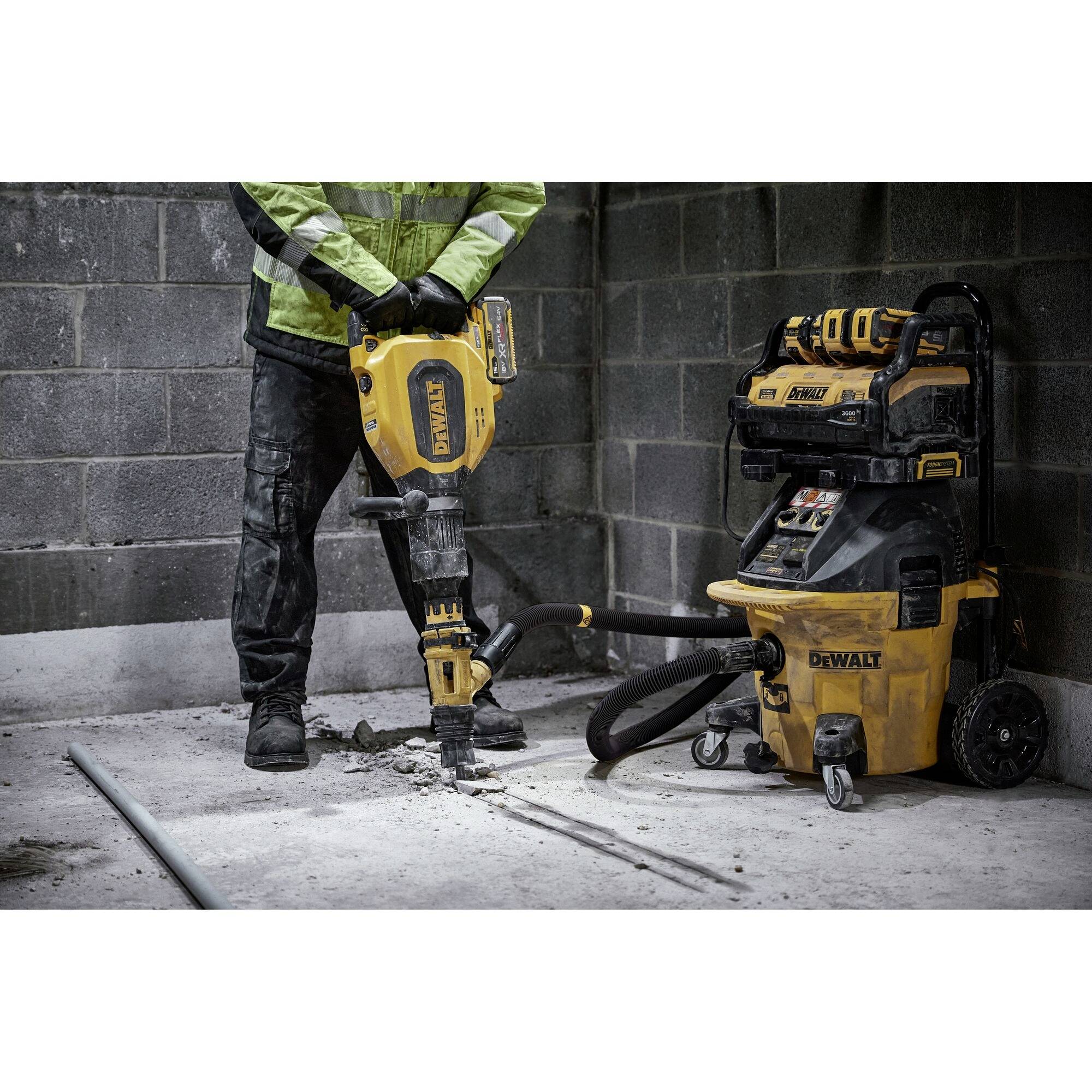 A person wearing work attire is using a demolition hammer on a concrete wall, connected to an industrial vacuum cleaner.