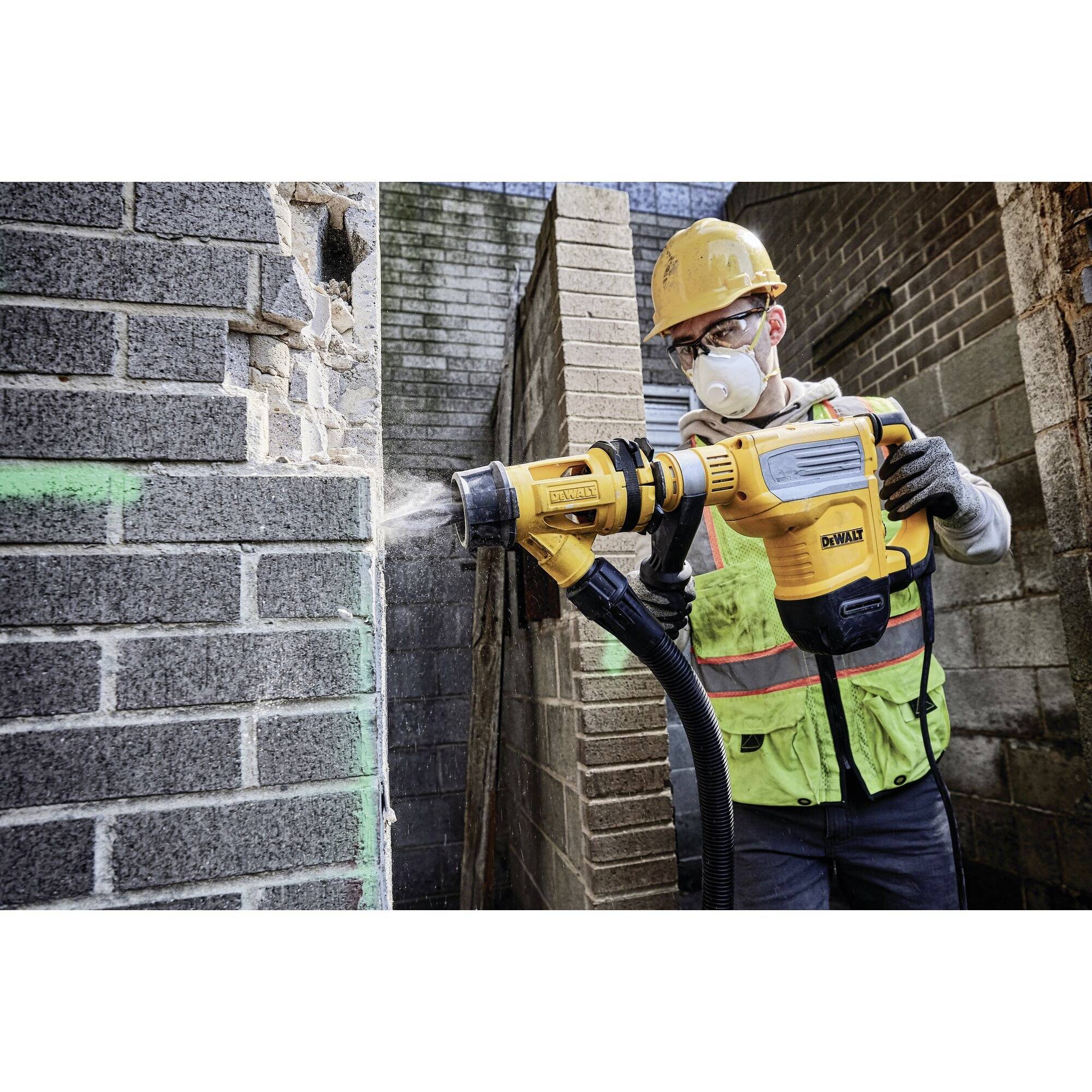 A person wearing safety glasses and a helmet is using a yellow drill hammer to break down a wall. Dust is visible.
