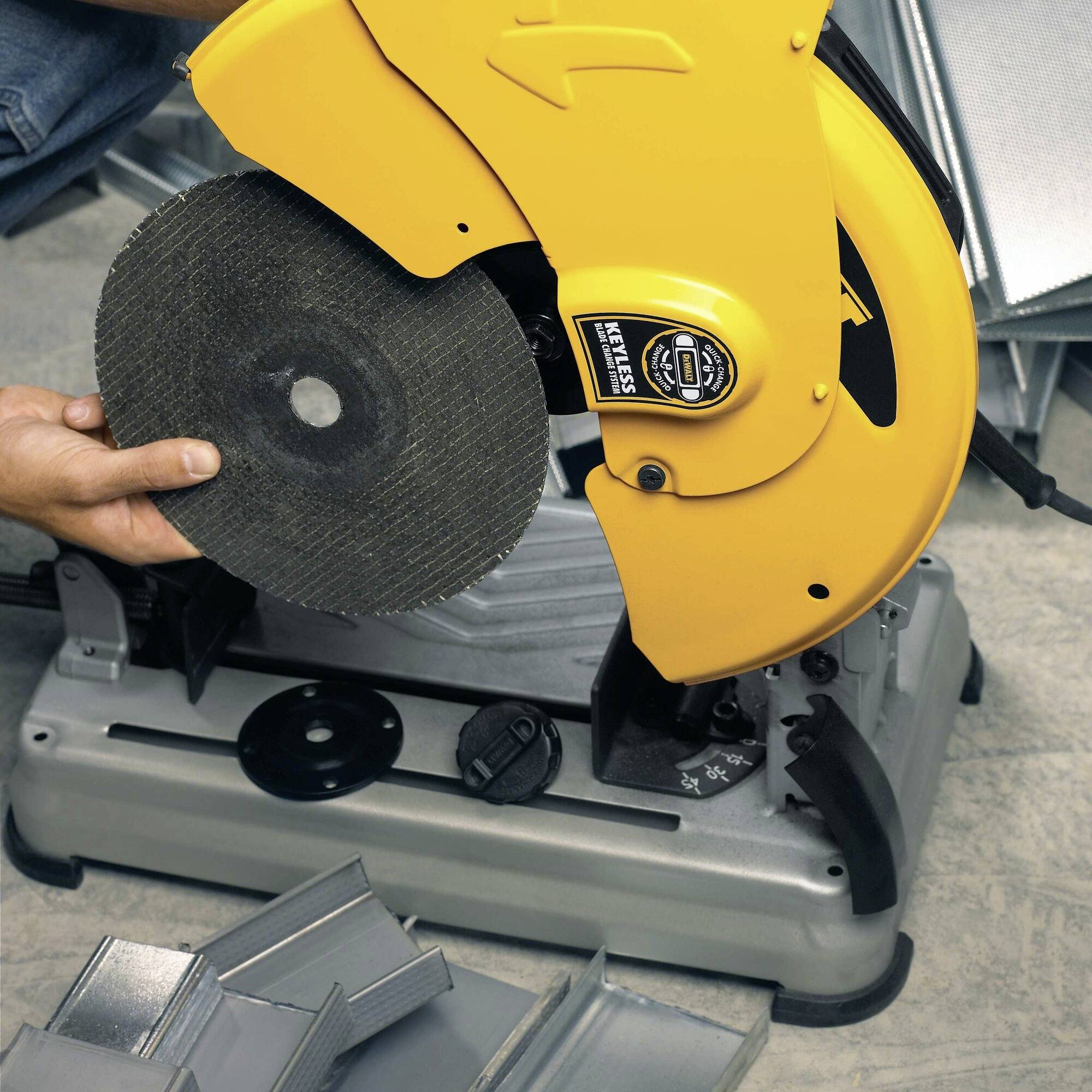 A yellow metal circular saw is being held by a person. It is cutting a piece of metal. Stacks of metal pieces can be seen in the background.