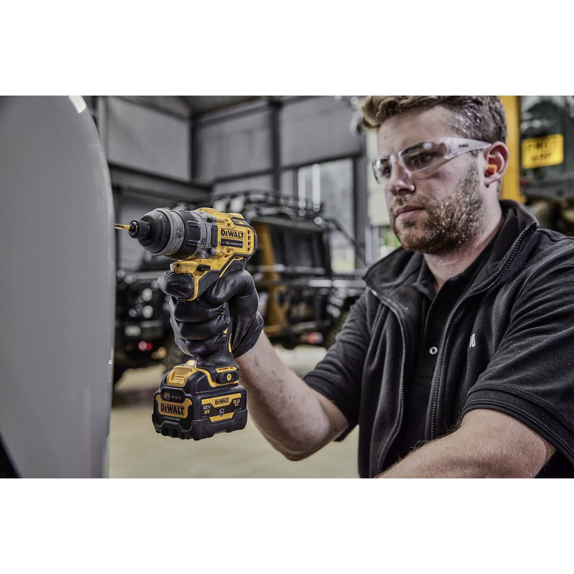 A man is using a yellow drill in a workshop. He is wearing safety glasses and appears focused while working.