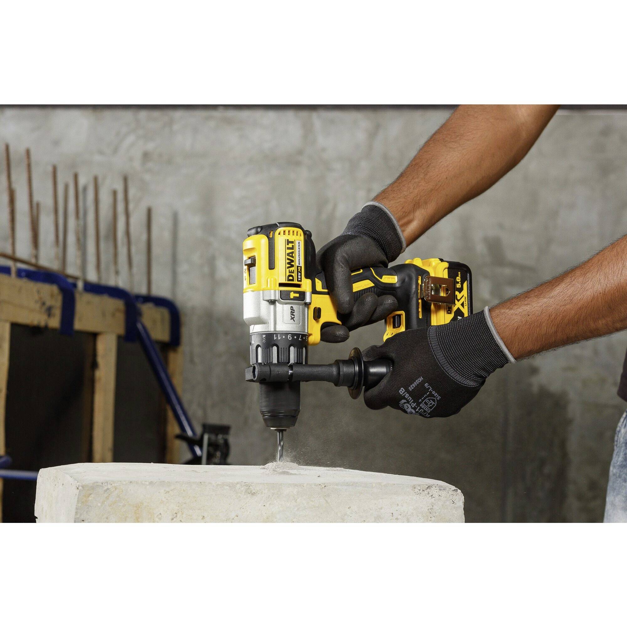 A person is drilling a hole in concrete using a yellow cordless drill. Construction tools and steel rods are visible in the background.