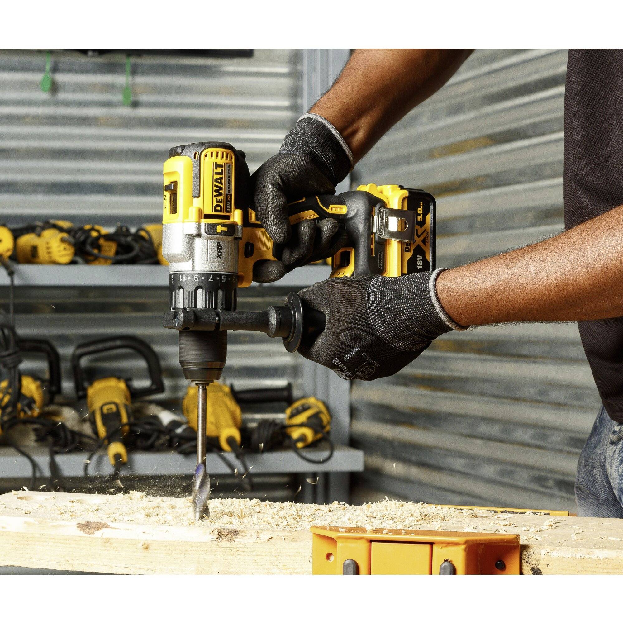 A person is drilling a hole in a wooden board using a yellow cordless drill. Various tools can be seen in the background.