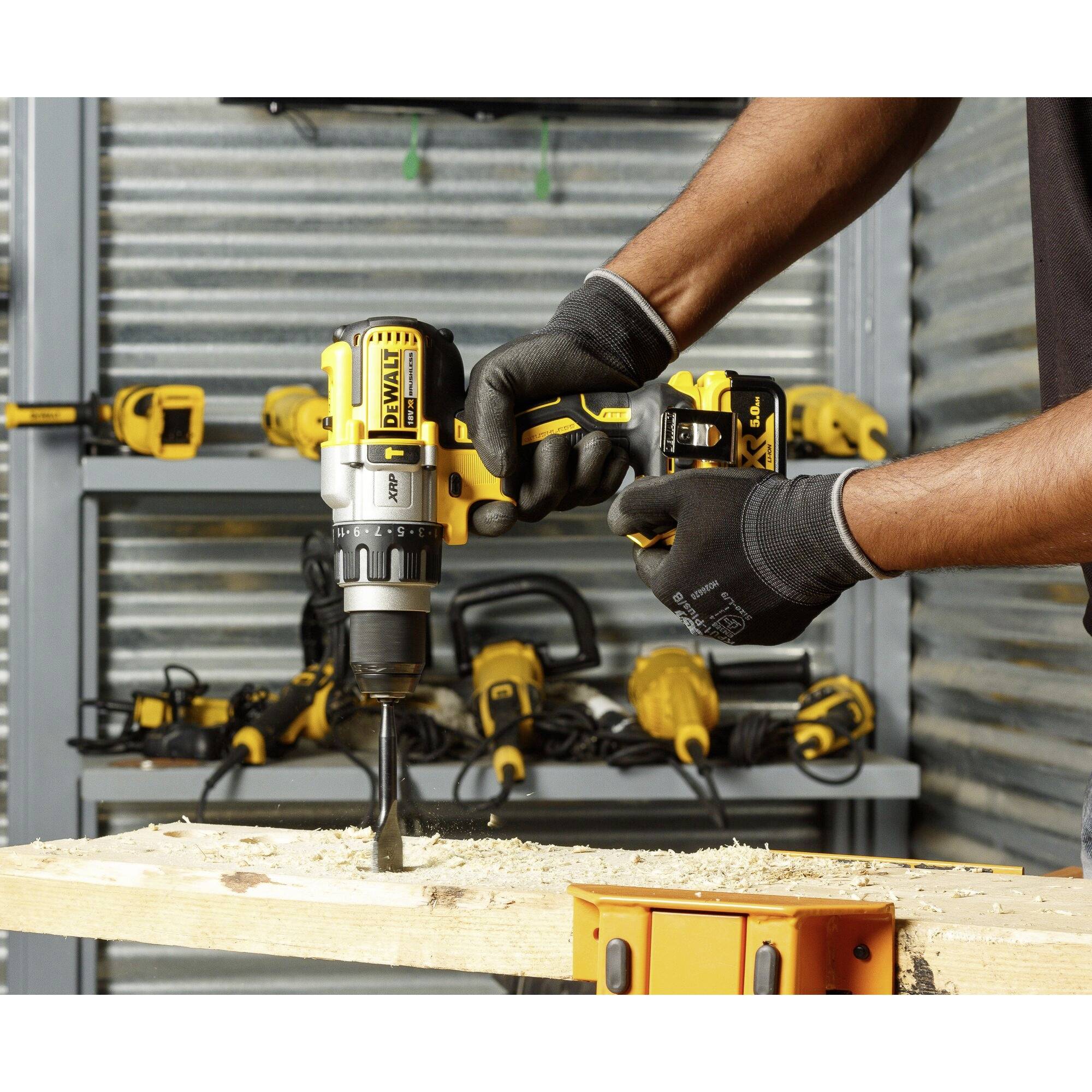 A person is drilling into a piece of wood with an electric drill, with further tools mounted on a metallic wall in the background.