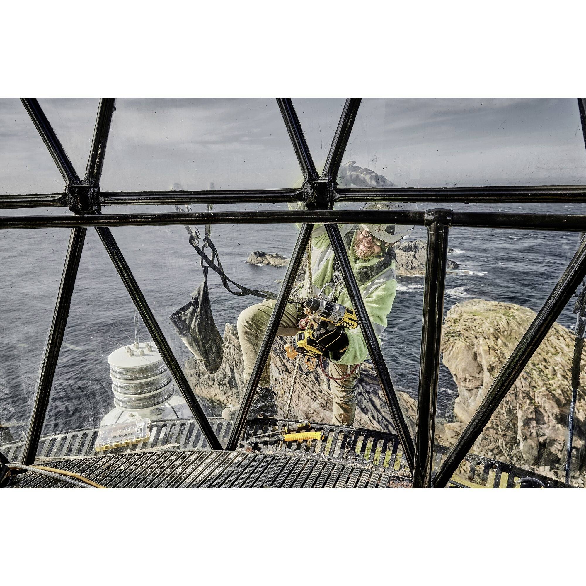 A person is cleaning the exterior of a lighthouse. The sea and rocks are visible in the background.