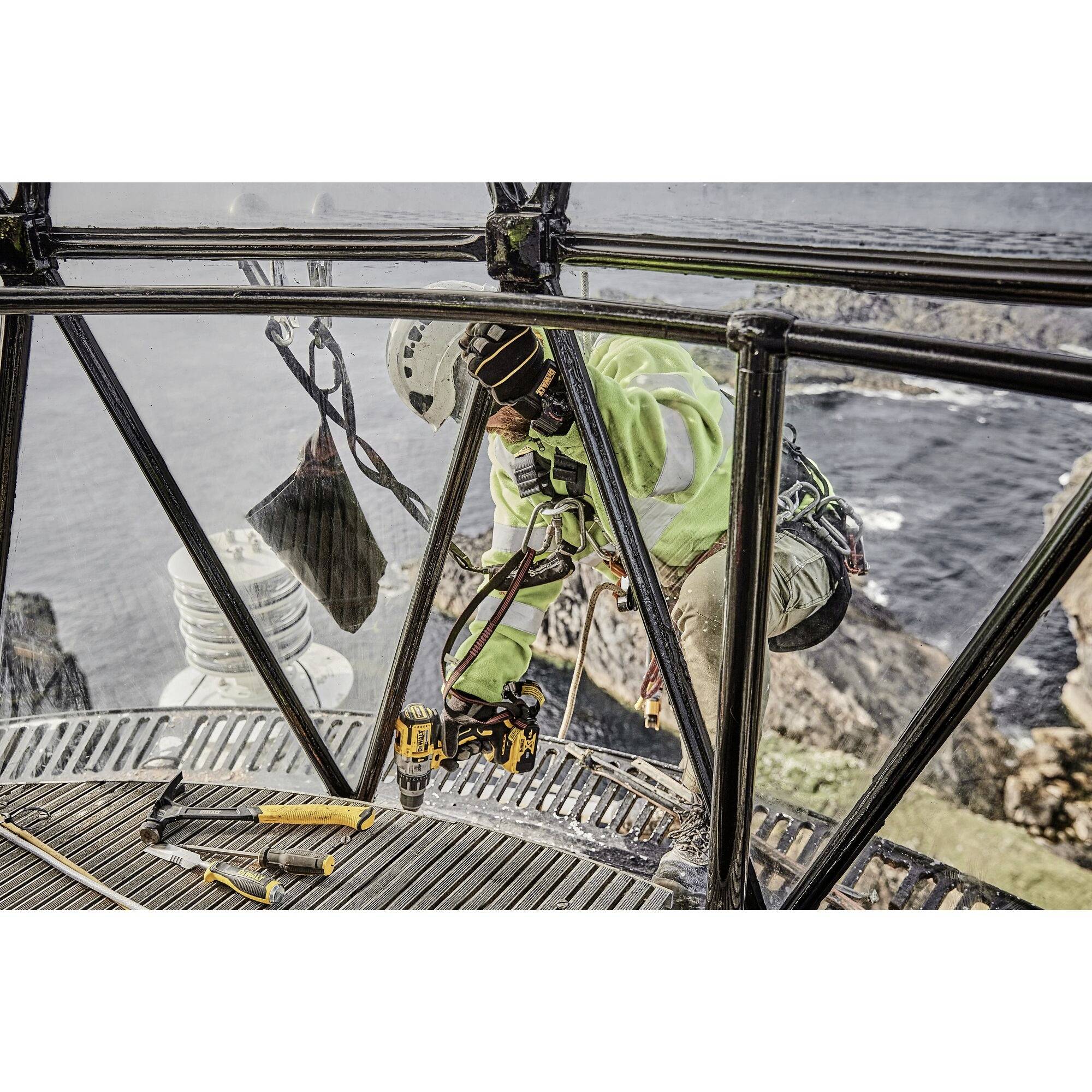 A person in safety clothing is working on the exterior of a lighthouse above a steep cliff edge, with tools nearby.