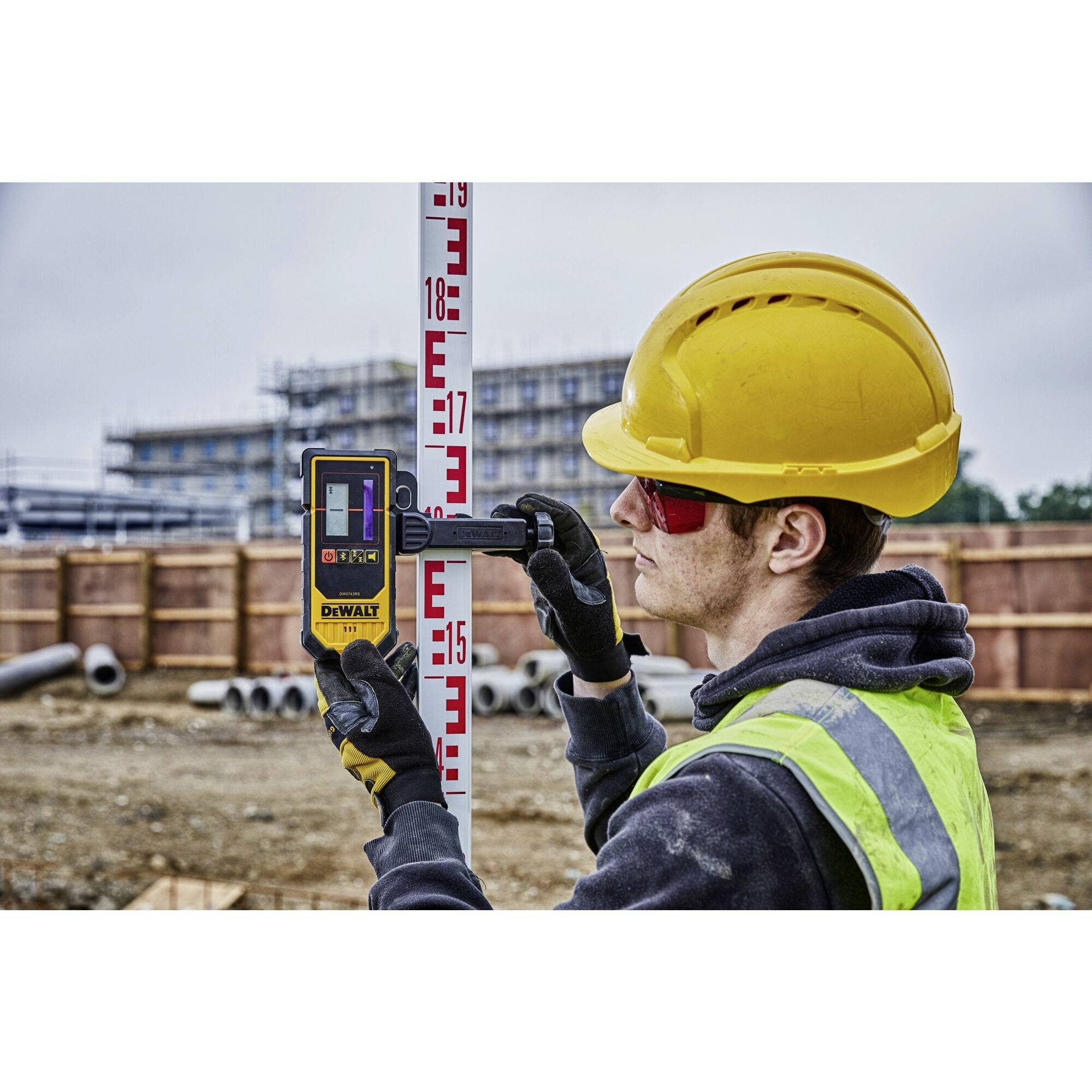 A construction worker in protective gear is measuring height on a building site using a laser distance meter against a measuring pole.