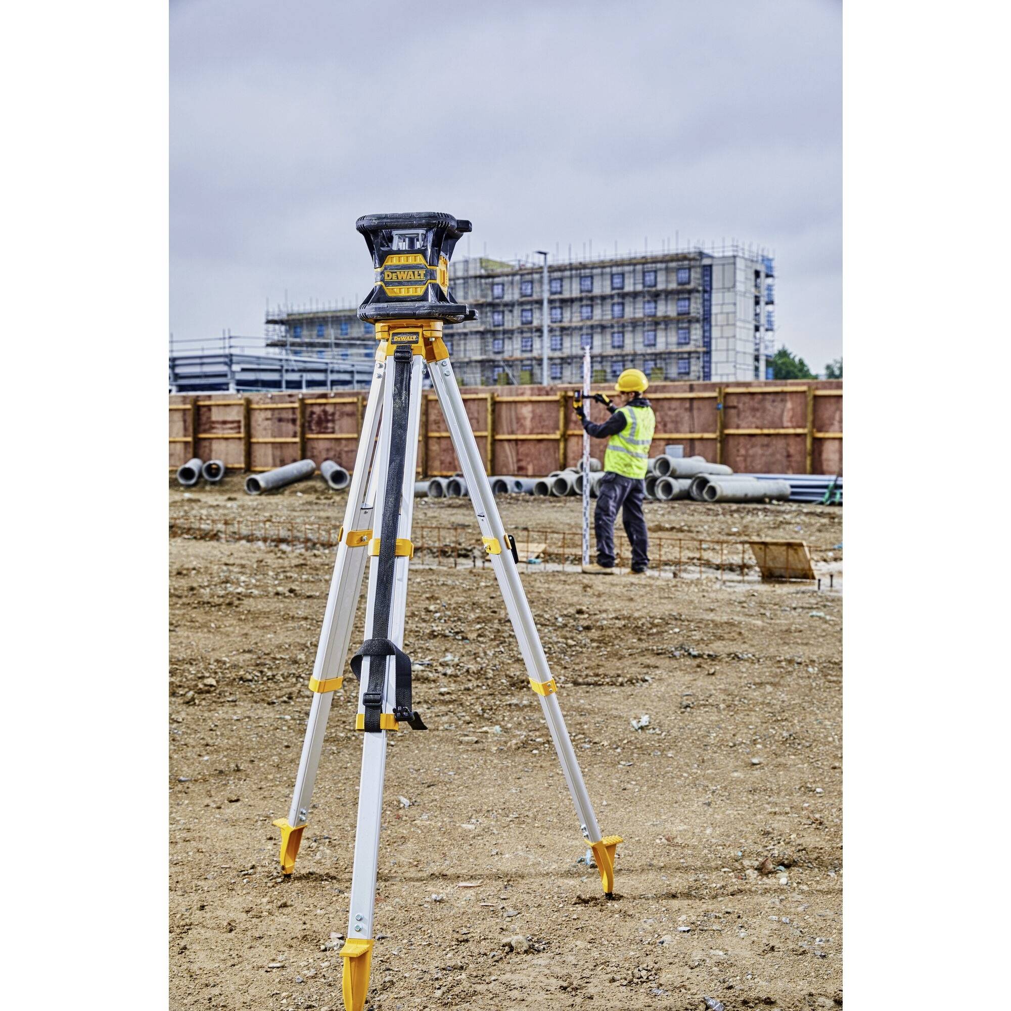 A construction site with a surveying instrument in the foreground. In the background, a person wearing protective clothing is working with pipes.