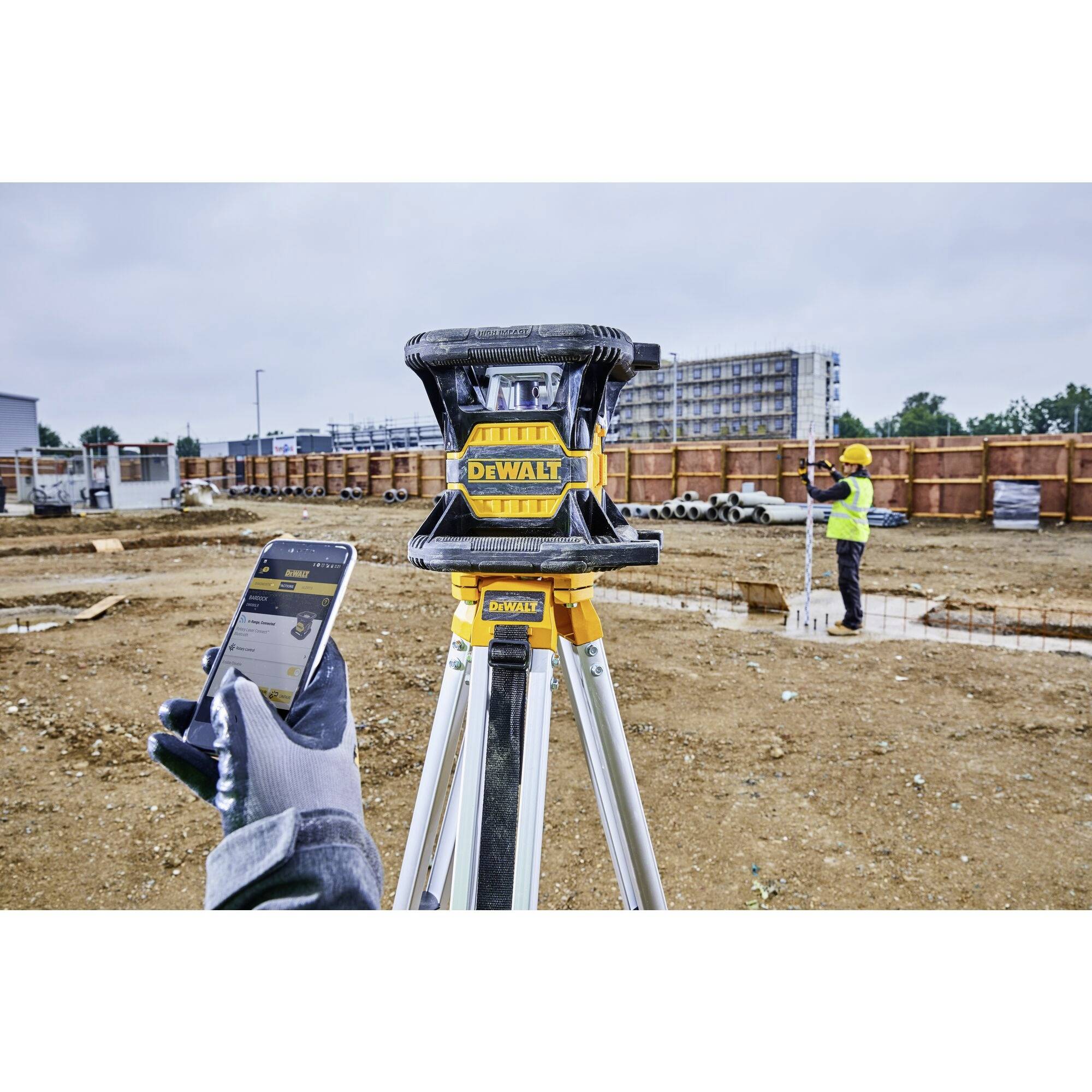 A construction worker is using a smartphone to remotely control a laser level on an outdoor construction site.