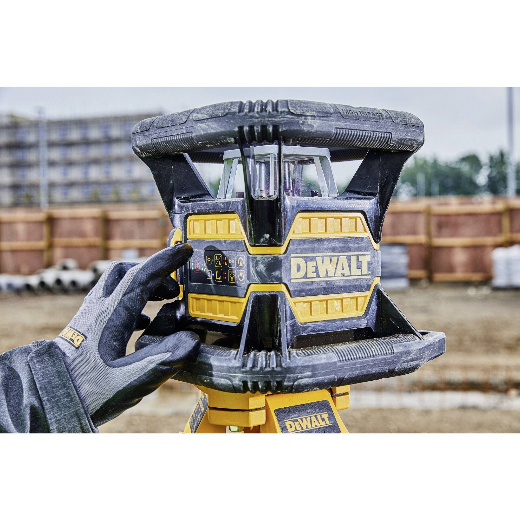 A person is operating a laser levelling device on a construction site. Buildings and materials are visible in the background.