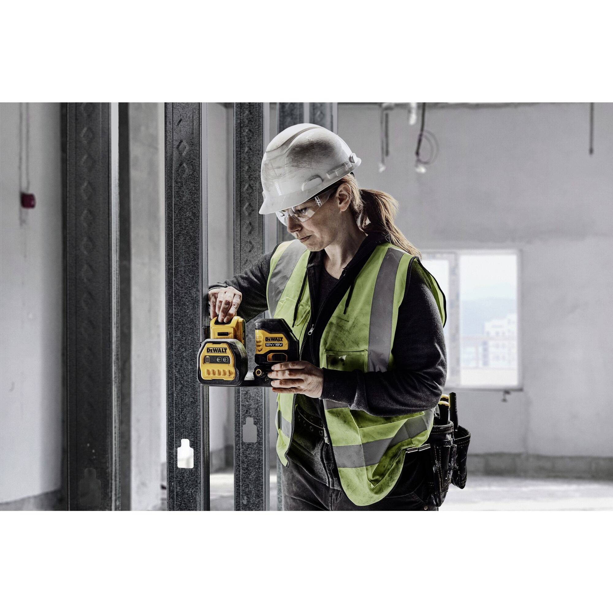 A female construction worker is using a laser levelling device on a construction site. She is wearing a white hard hat and a yellow high-visibility vest.