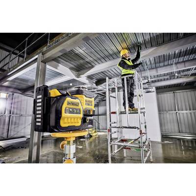 A construction worker on scaffolding is installing a ceiling panel in an industrial hall, with a laser levelling device visible in the foreground.