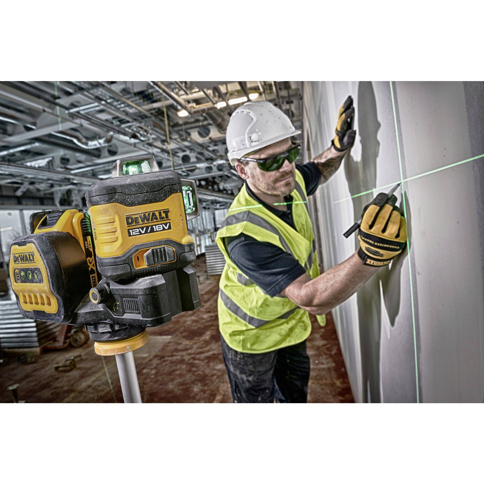 A construction worker wearing a hard hat and safety glasses is using a green laser measuring device on a wall. He is wearing a high-visibility yellow vest.