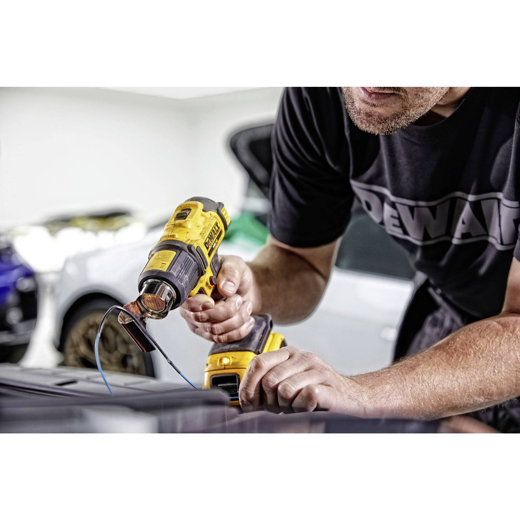 A person is using a cordless drill to fasten a car part in a workshop. Battery and cables are visible.