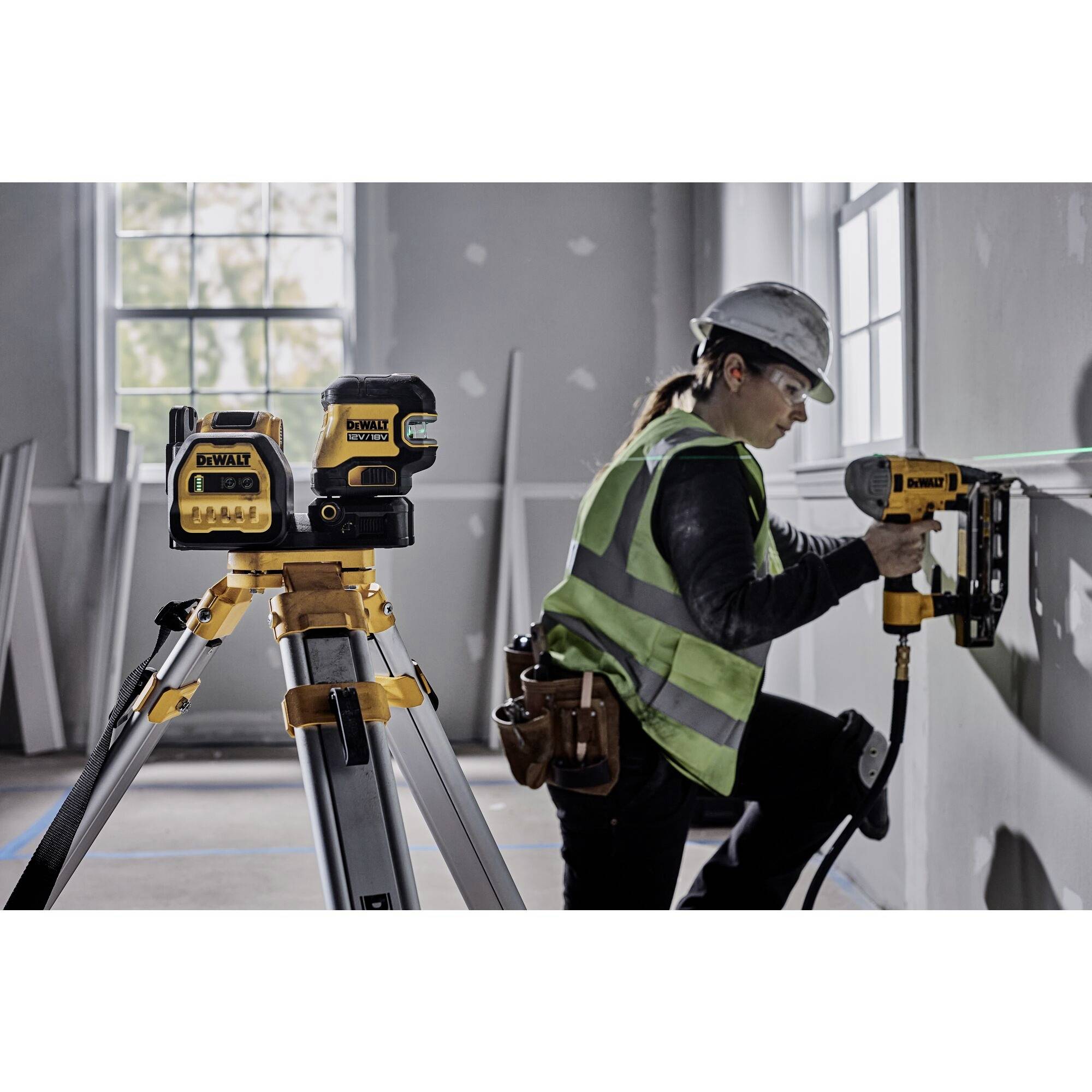 A person wearing protective clothing is working with a laser levelling device mounted on a tripod. Windows can be seen in the background.