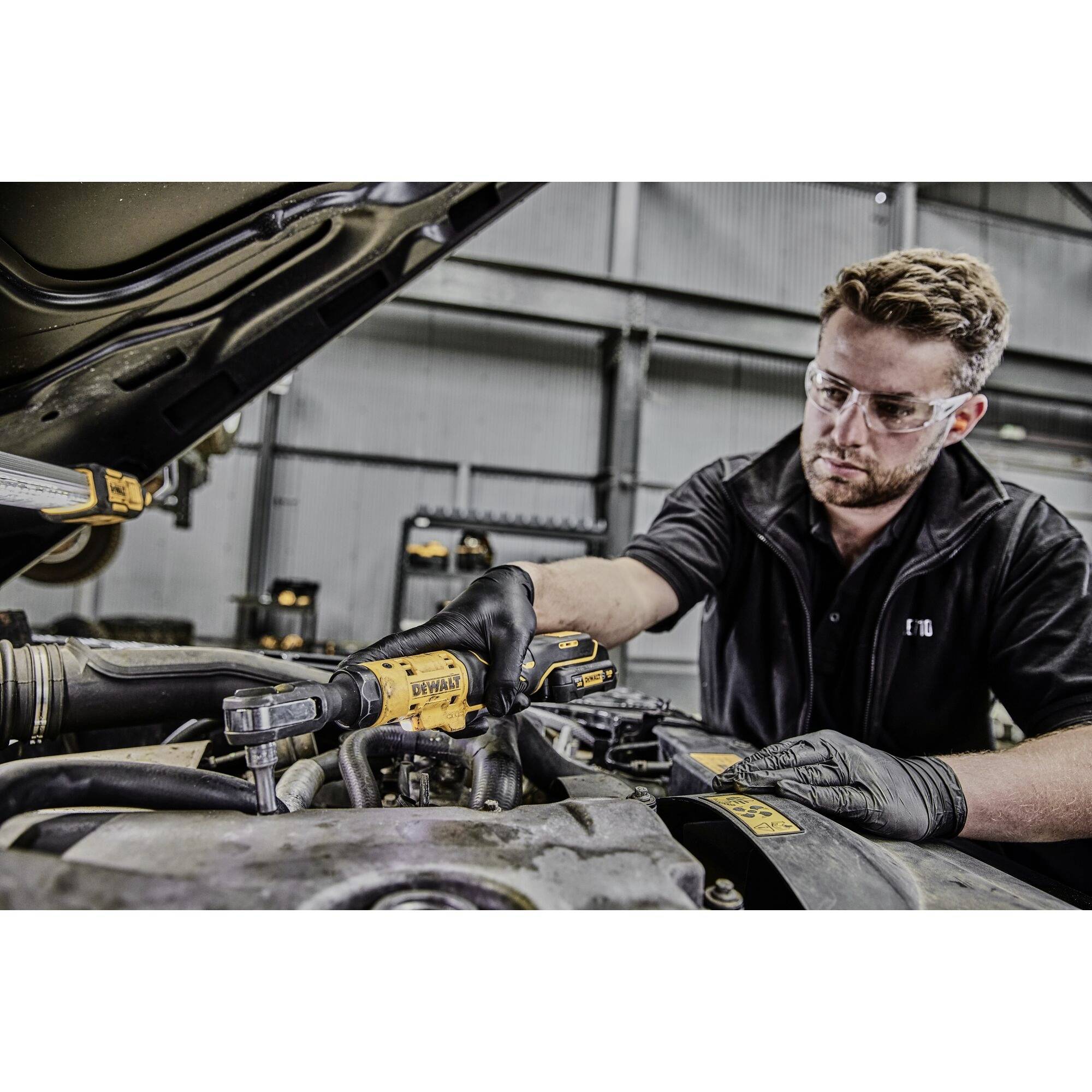 A mechanic is working in a workshop with an electric tool under the open bonnet of a vehicle. He is wearing safety glasses and gloves.