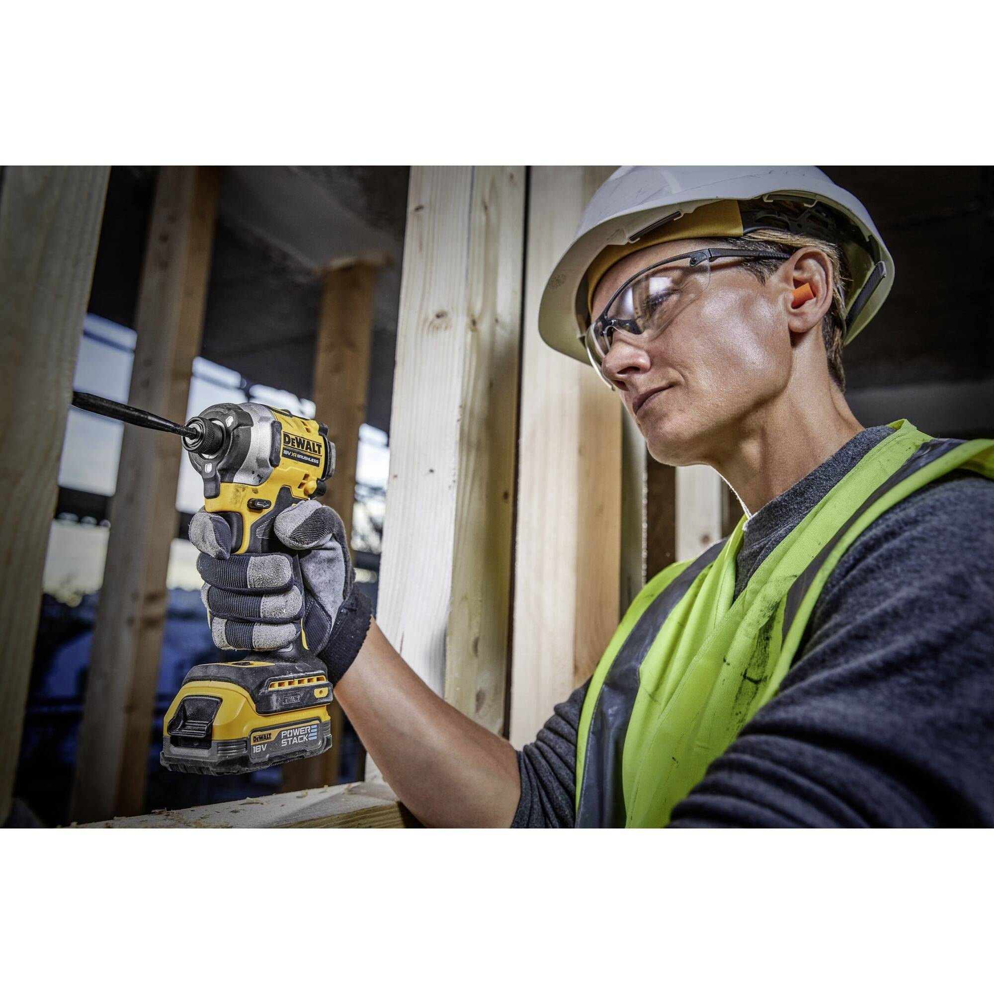 A construction worker wearing safety glasses and a hard hat is using a cordless drill to connect wooden boards at a building site.