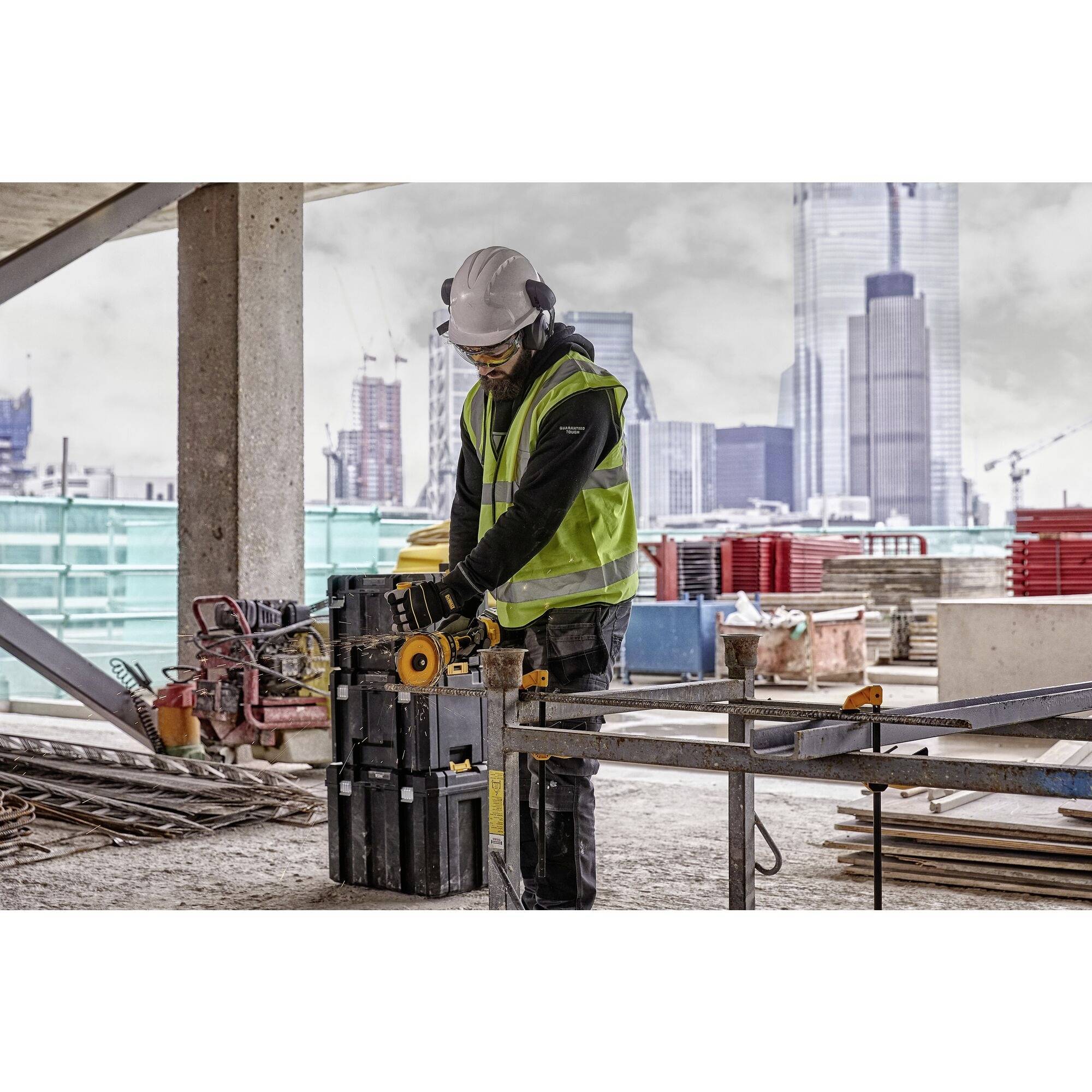 A construction worker wearing a hard hat and safety glasses is cutting a piece of metal on a building site. Skyscrapers can be seen in the background.