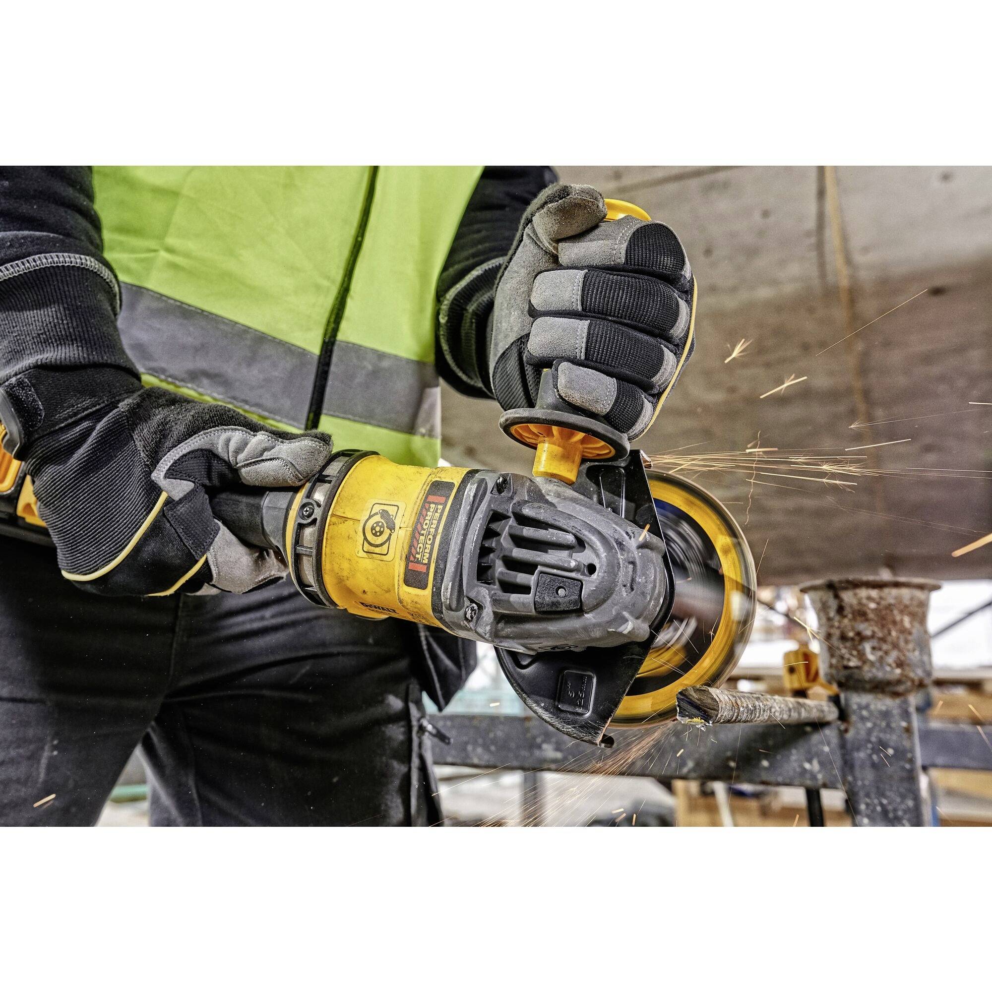 A person in work attire is using an angle grinder, with sparks flying. Safety clothing is being worn, with a workbench in the background.