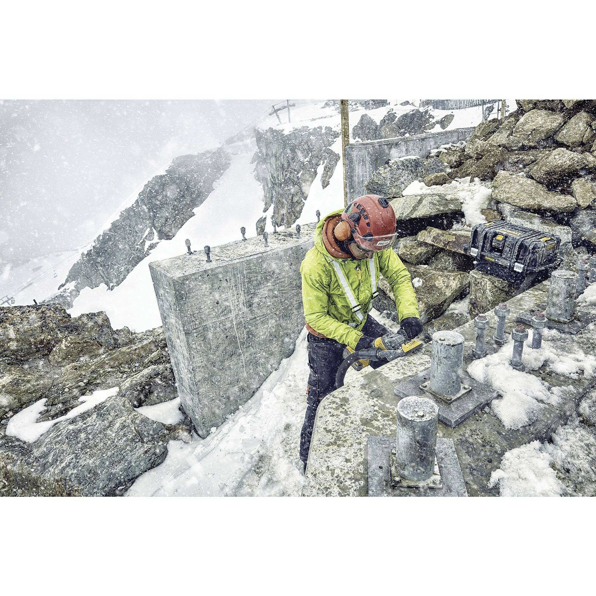 A person in high-visibility clothing and a helmet is working on metal structures in the mountains during snowfall. Tools and equipment are visible.