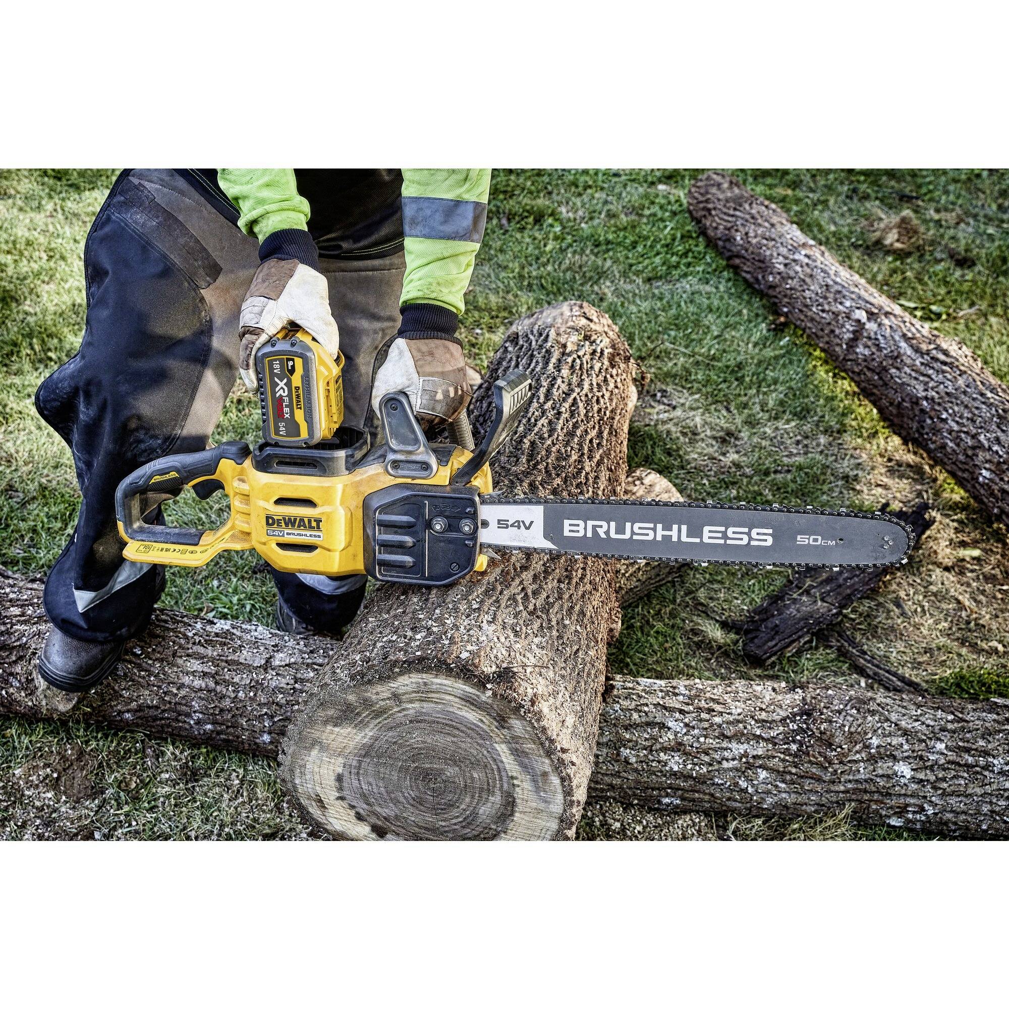 A person is sawing a tree trunk with a yellow cordless chainsaw. The lawn is visible in the background.
