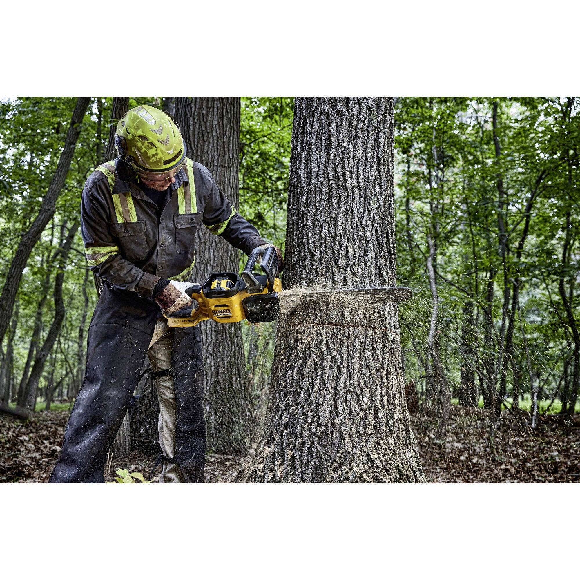 A person in protective clothing is felling a tree using a chainsaw in a wooded area. Wood chips are flying, and the tree is half-cut.