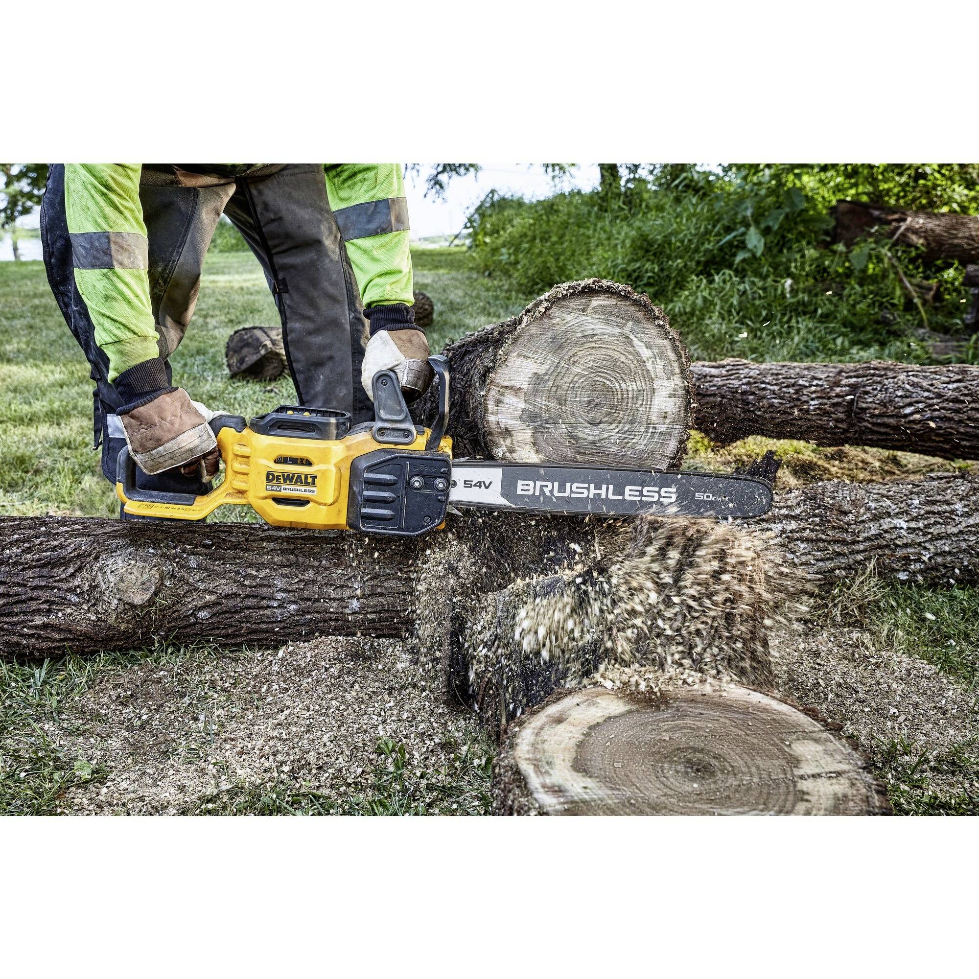 A person wearing protective clothing is sawing a tree trunk outdoors with a yellow chainsaw. Sawdust is flying through the air.