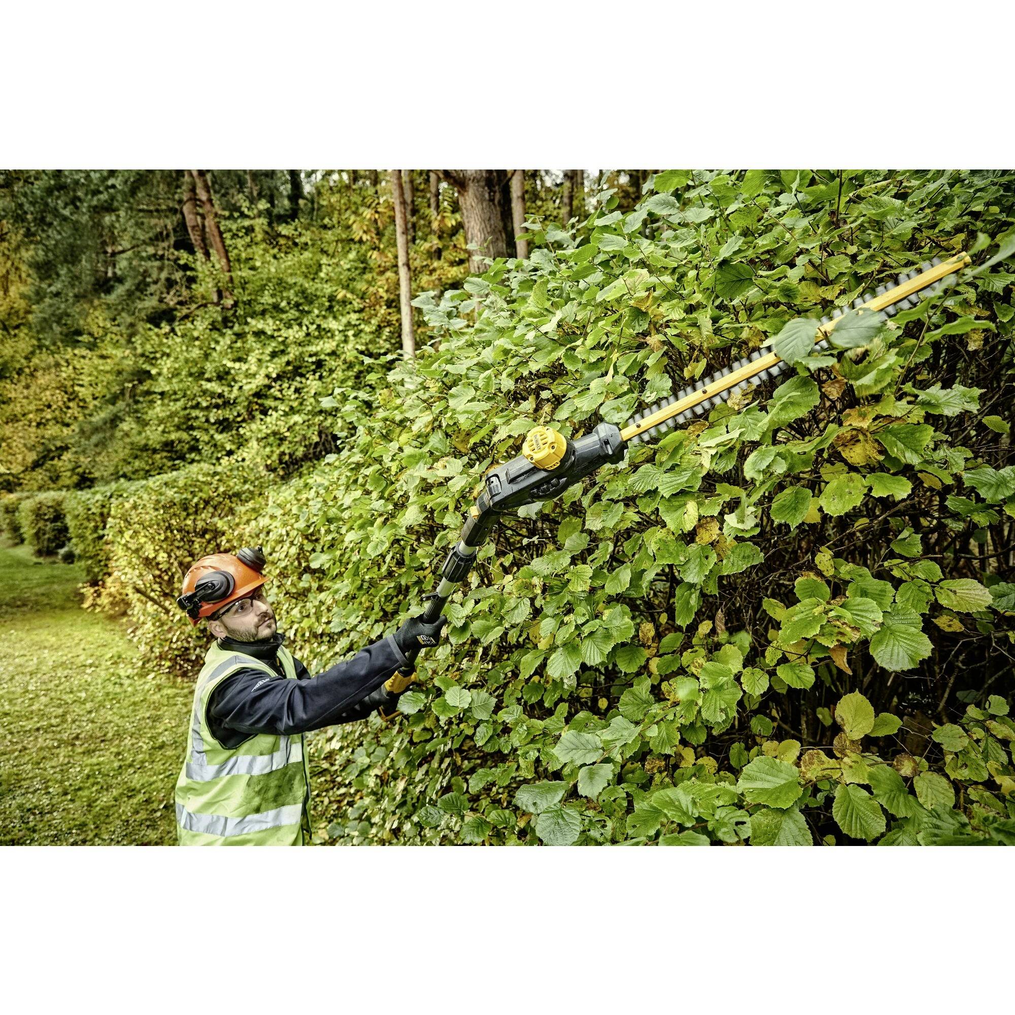 A man is trimming a tall hedge of green foliage with an electric hedge trimmer. He is wearing protective clothing and a helmet.