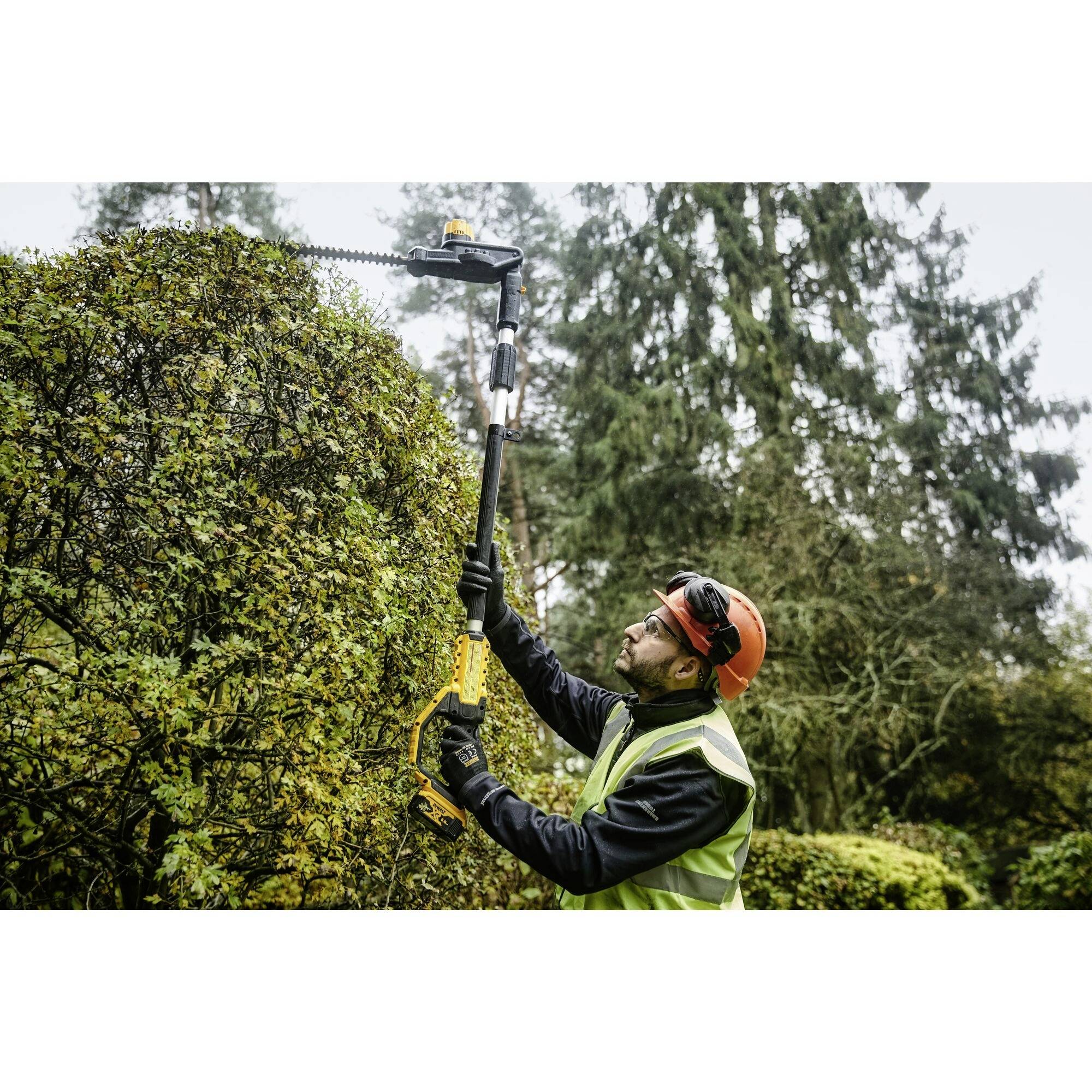 A person wearing a protective helmet and vest is using a telescopic hedge trimmer to cut a tall hedge in the garden.