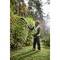 A worker in safety clothing is using an electric hedge trimmer to cut a tall hedge in a woodland area.