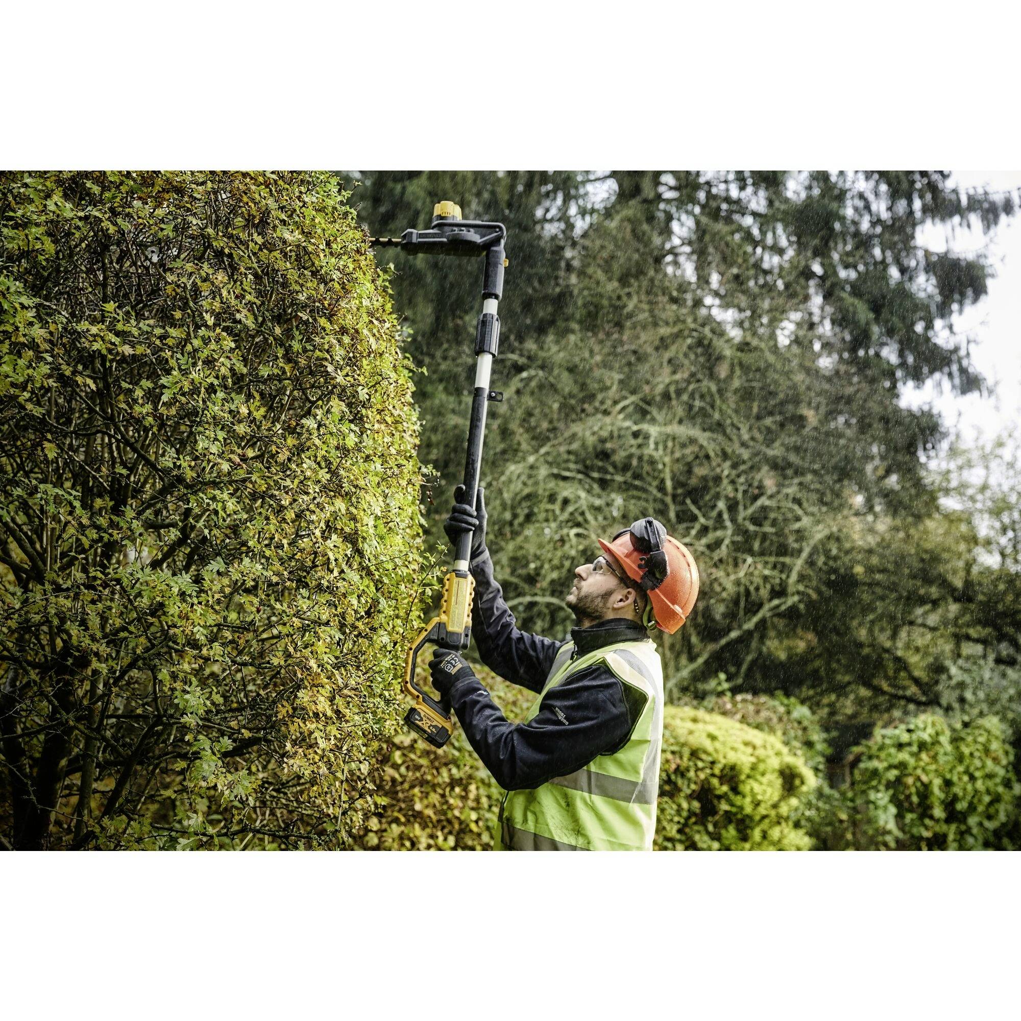 A gardener wearing a safety helmet and hi-vis vest is trimming a tall hedge with an electric hedge trimmer in a garden.