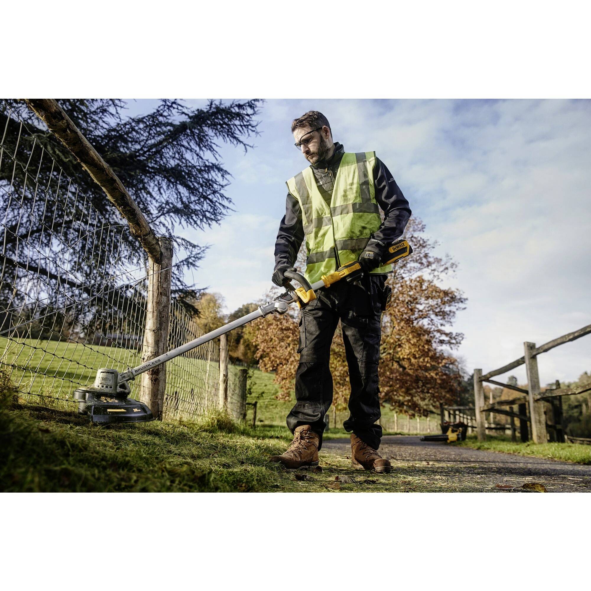 A man in work attire and a high-visibility vest is using a strimmer along a fence on a sunny path beside a meadow.