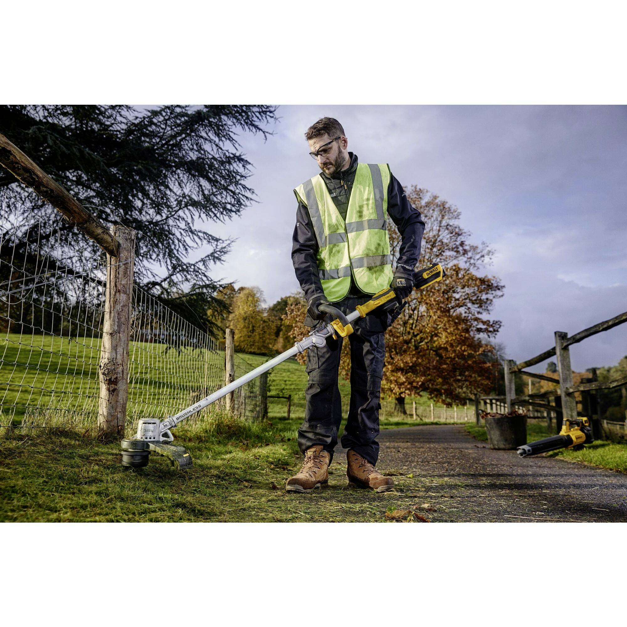 A person wearing a high-visibility safety vest and protective glasses, operating a strimmer along a fence beside a rural road, with autumn trees in the background.