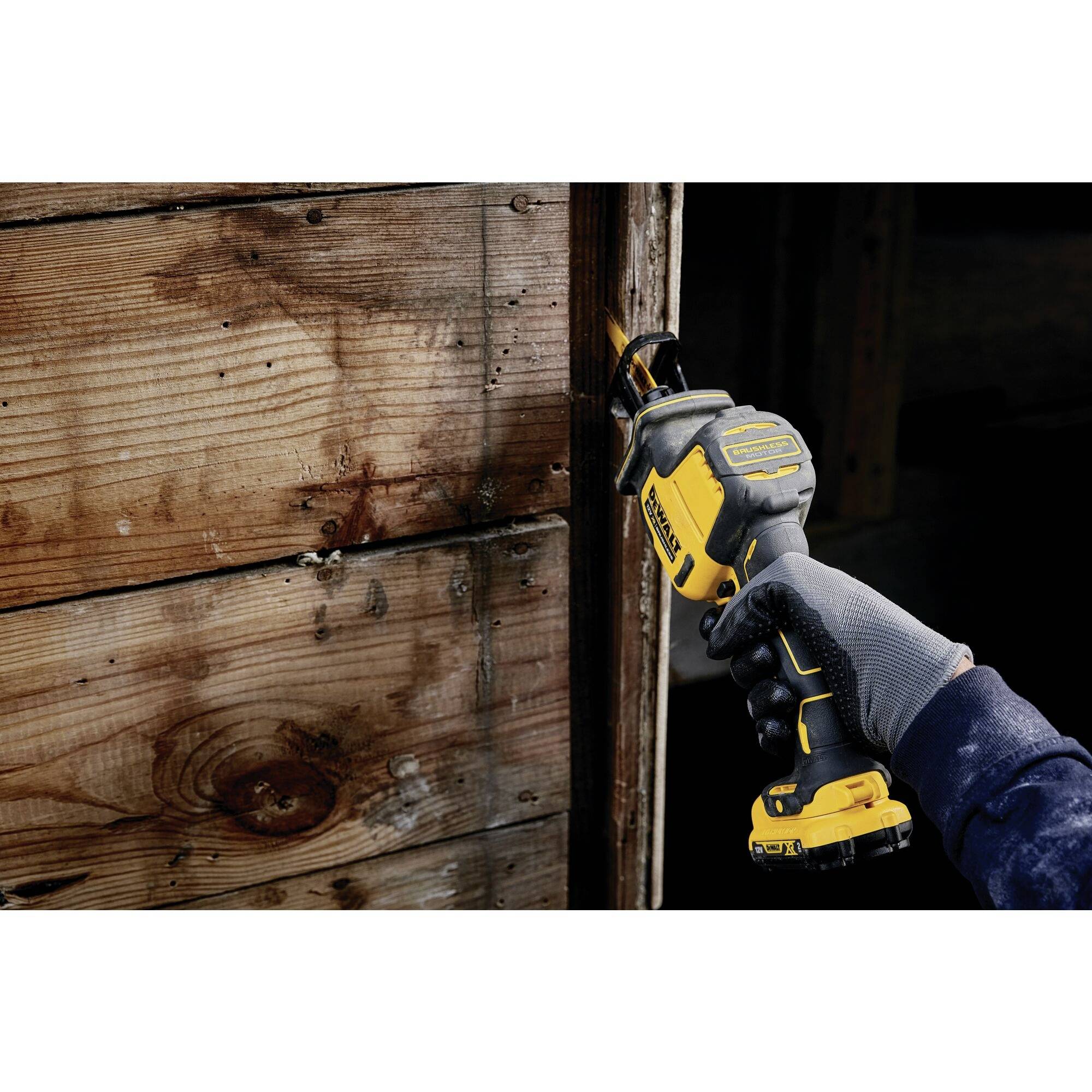 A person is sawing through a wooden board with a yellow, cordless jigsaw. The board shows visible wood grain and signs of wear.