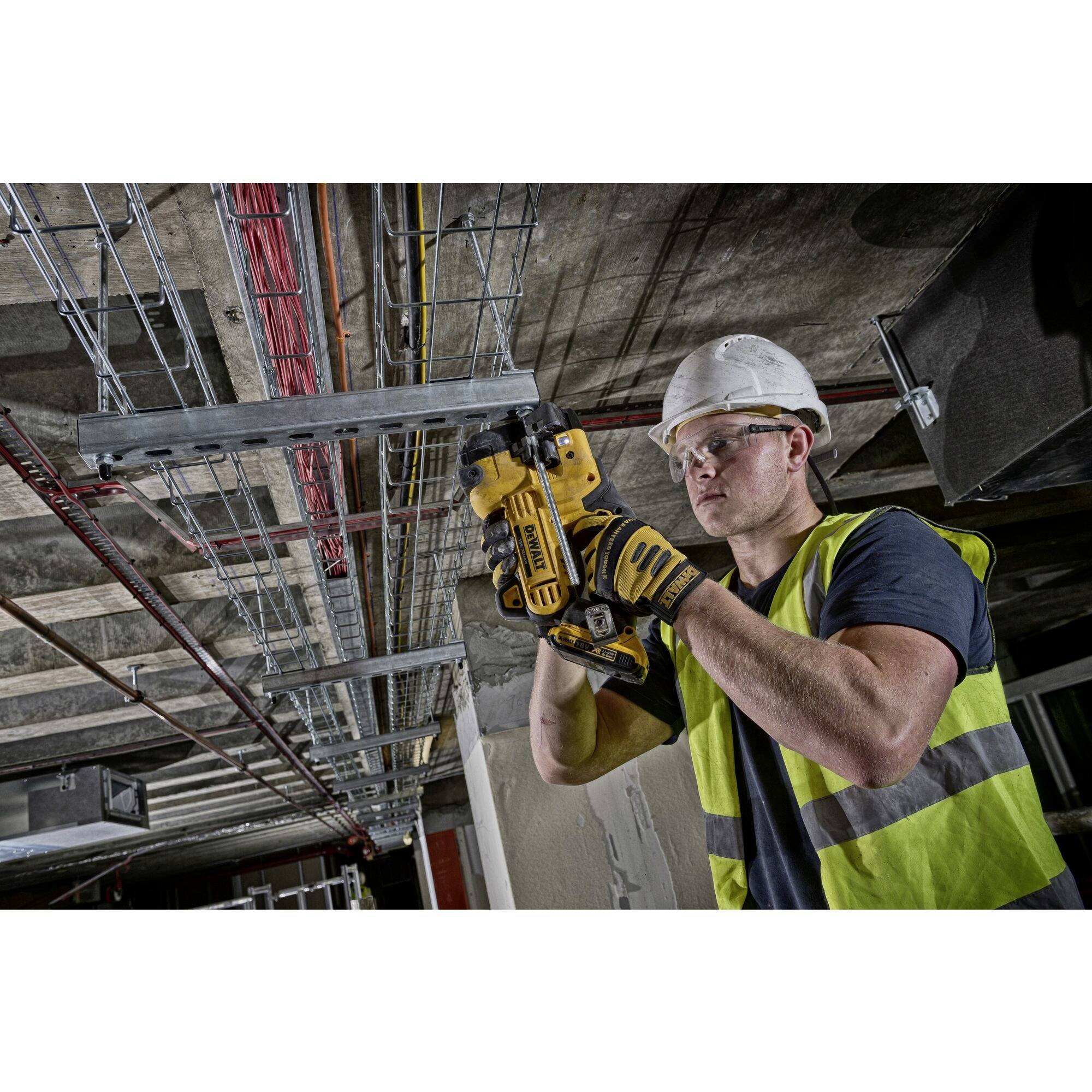 A worker wearing a hard hat and high-visibility vest is installing cable conduits on the ceiling using a tool. Construction area inside a building.