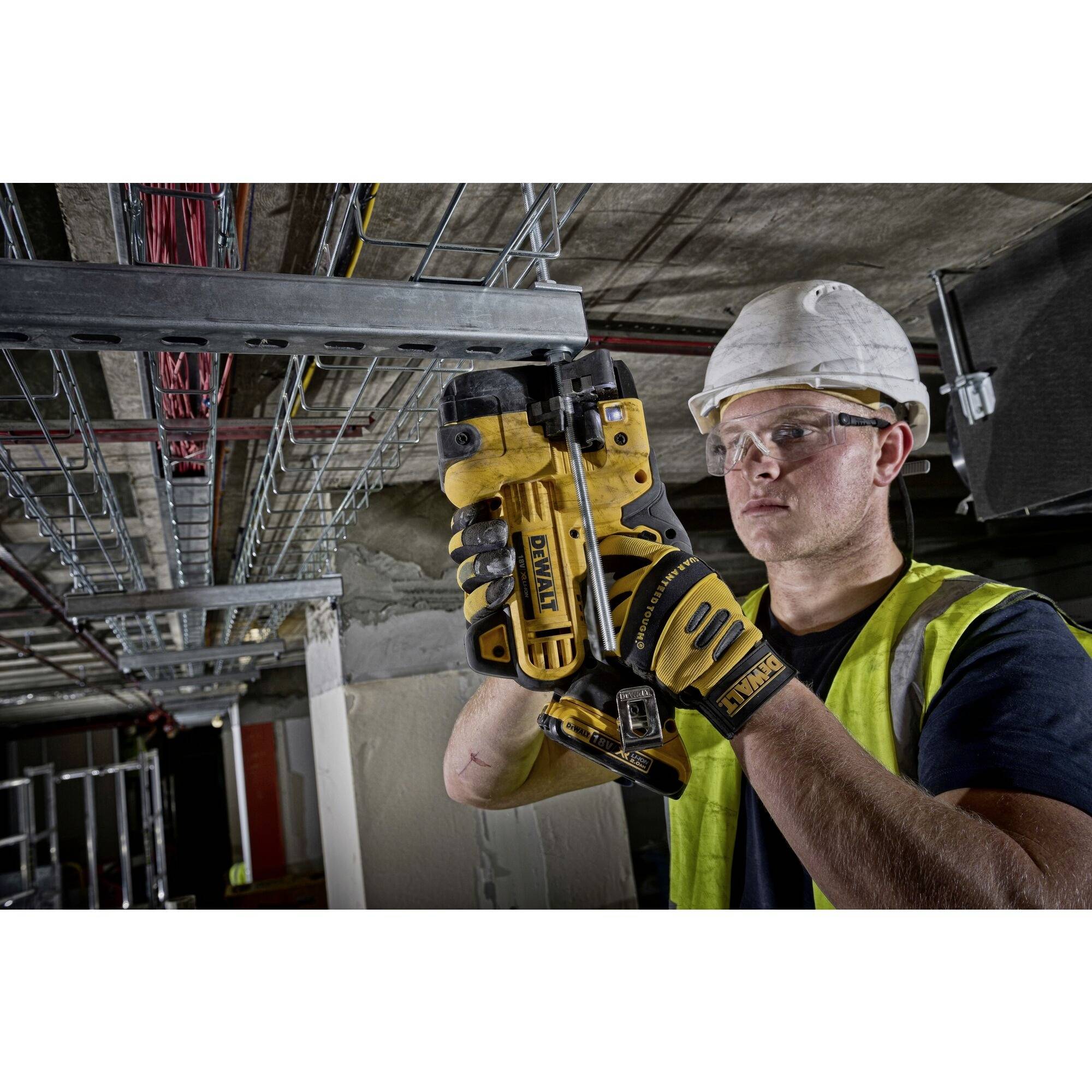 A worker wearing a hard hat and safety glasses is securing cables with a yellow tool to a metal structure inside a building.