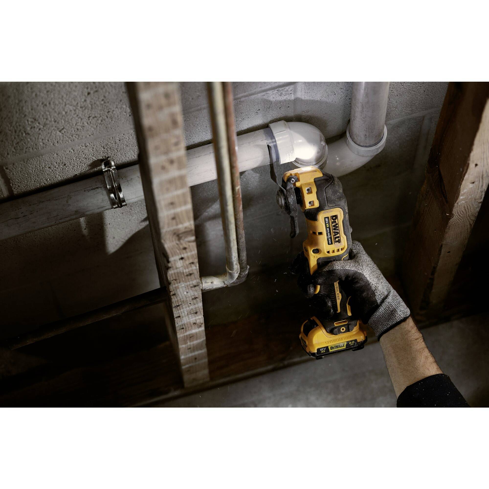 A person is using a cordless drill driver to secure a pipe in a basement area. The tool and hand are in focus.