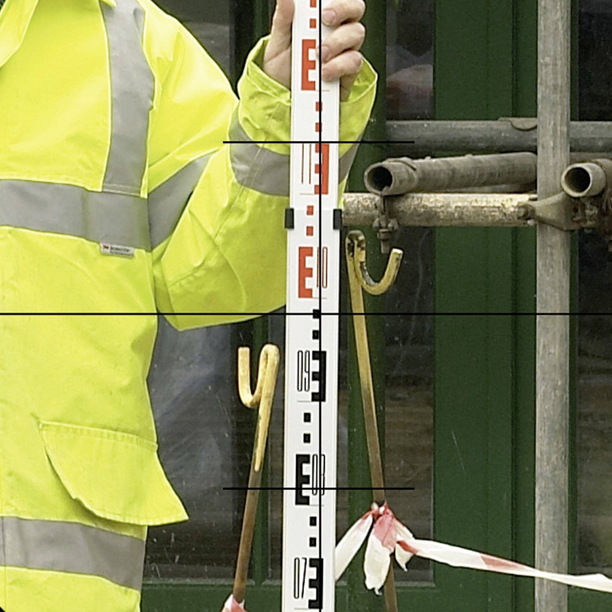 A person wearing high-visibility clothing is holding a levelling staff in front of a scaffolding. The scene suggests construction or surveying work.