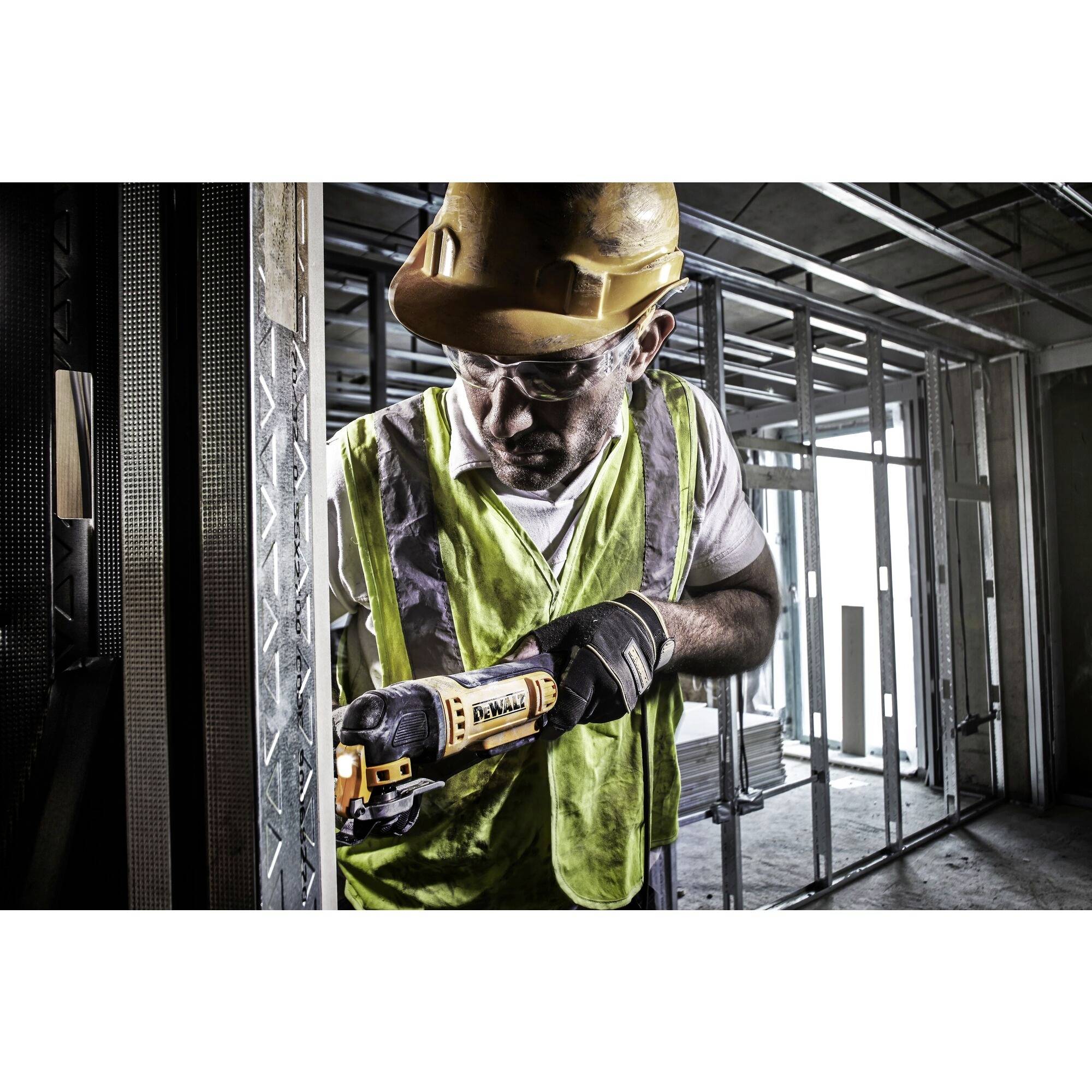 A construction worker in protective clothing and hard hat is working with an electric tool on a metal frame within a building scaffolding.