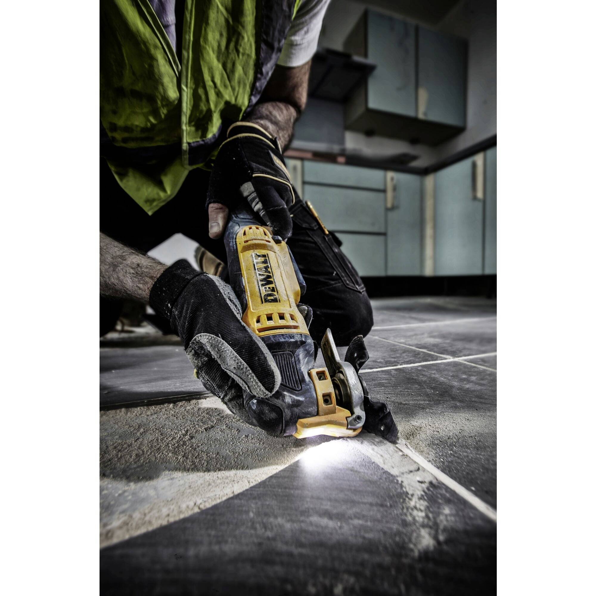 A worker wearing protective gloves uses an electric tool to cut tiles in a kitchen. Dust and light are visible.