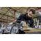 A man wearing safety glasses is working with an electric saw in a workshop. He is cutting a wooden board on a workbench.
