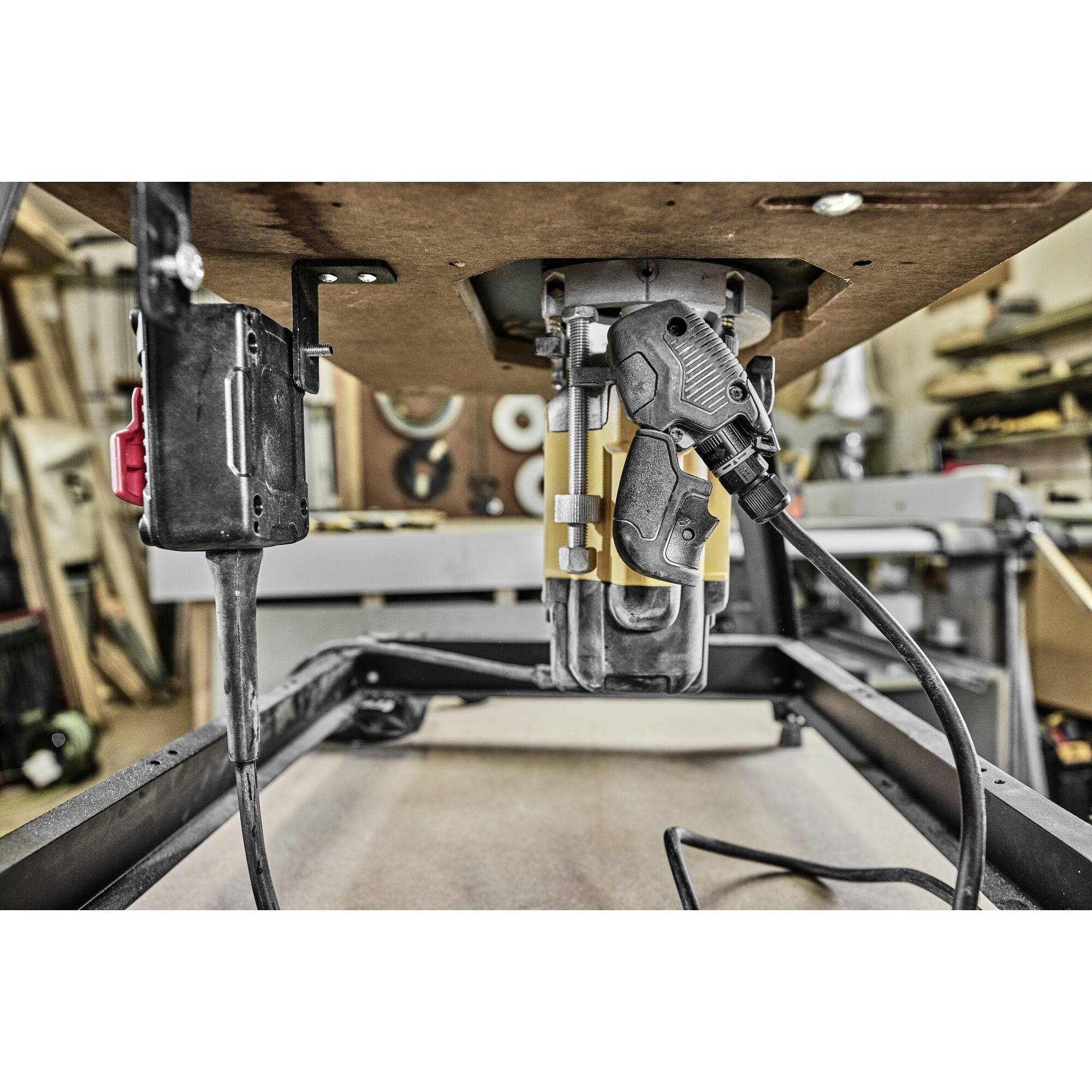 A workshop with a table router in the foreground. The router is mounted underneath a workbench, surrounded by various tools and woodwork in the background.