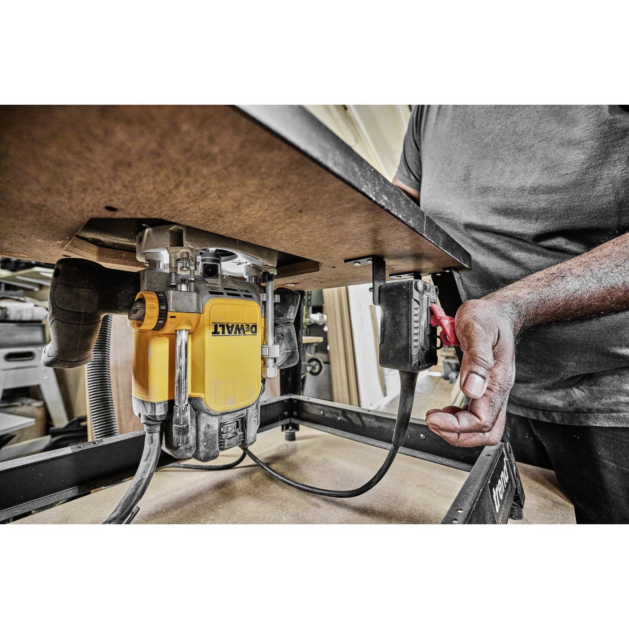 A person is working with a yellow table router in a workshop environment. The hand is reaching for the switch to adjust settings.
