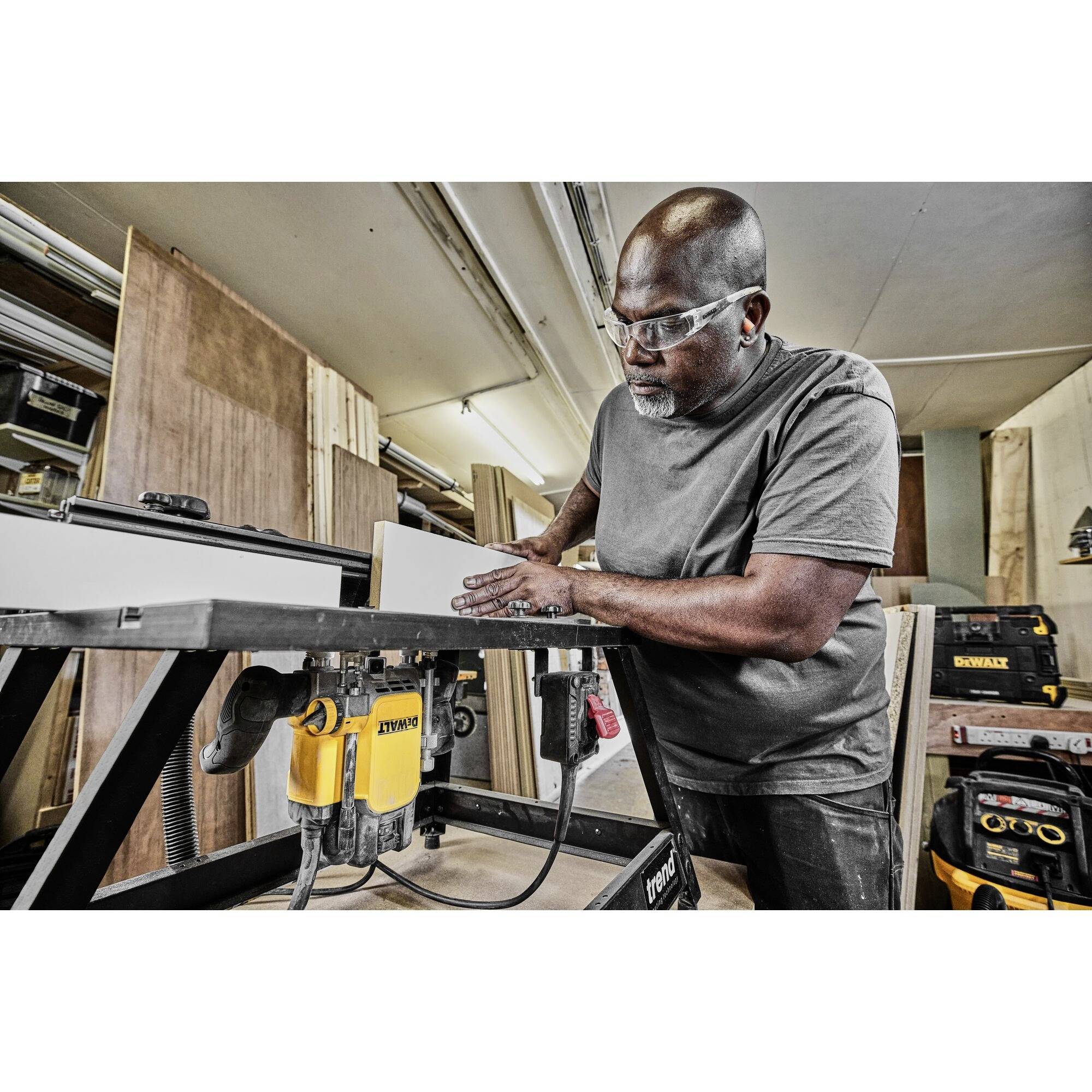A man is working intently on a table saw in a workshop, wearing safety glasses, surrounded by wood and tools.